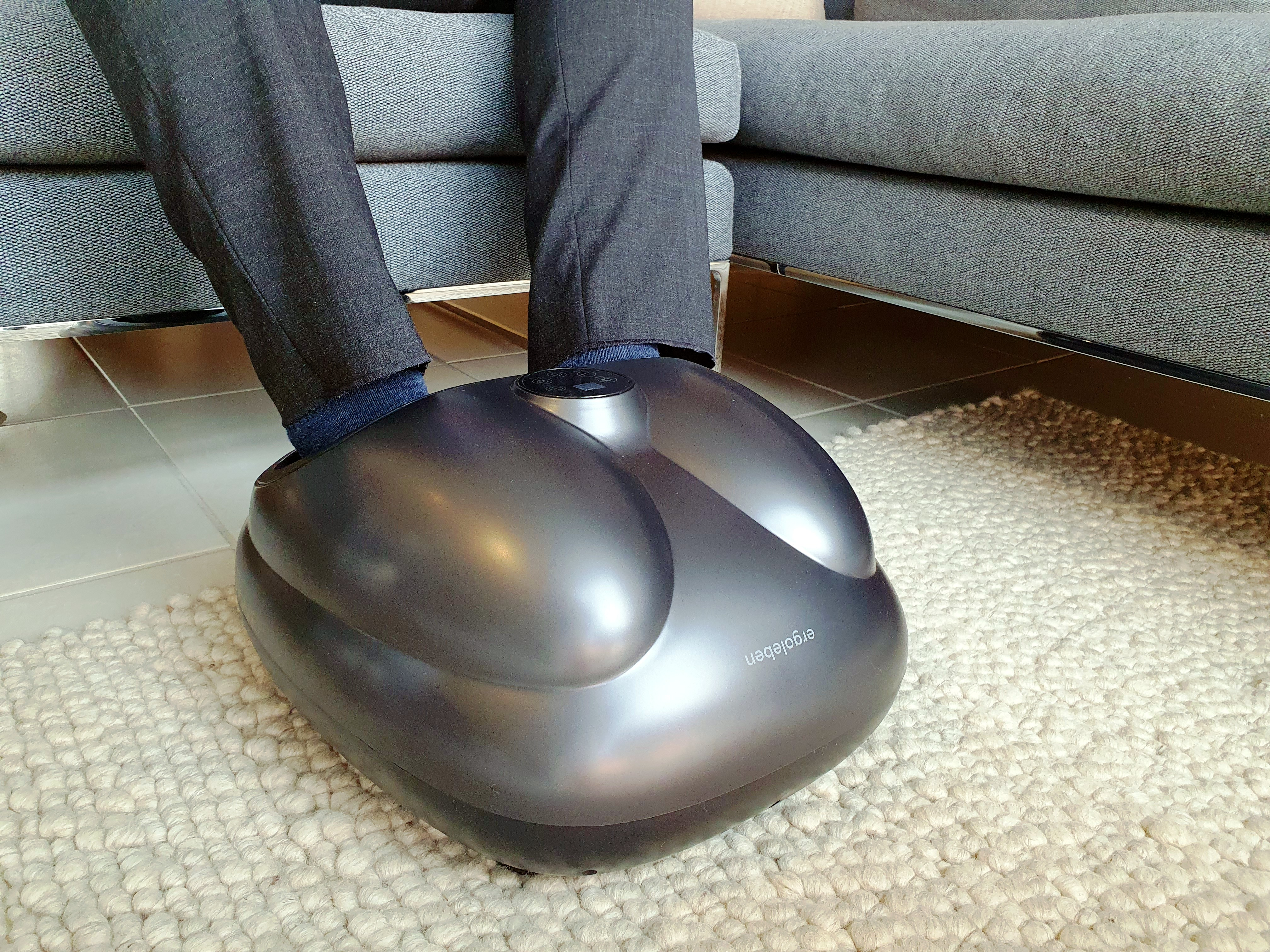 Füße einer Person in einem Fußmassagegerät auf einem Teppich, graues Sofa im Hintergrund.