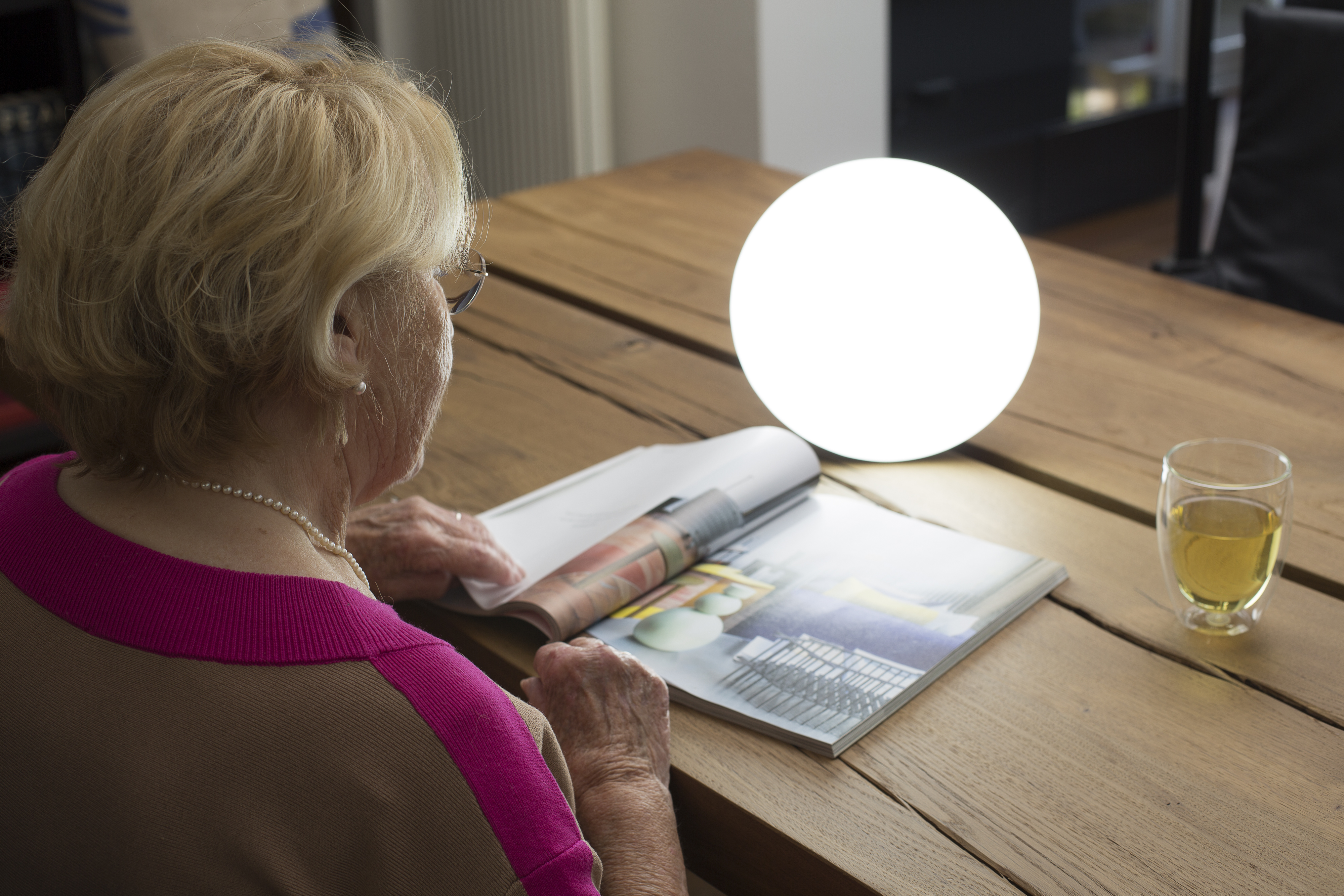 An elderly woman reads a magazine. A bright lamp and a glass of liquid sit on the wooden table.