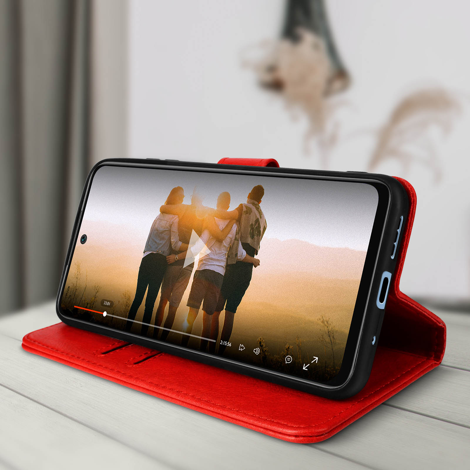 Een smartphone in een rode hoes. Het scherm toont een video van mensen. Achtergrond: houten tafel.