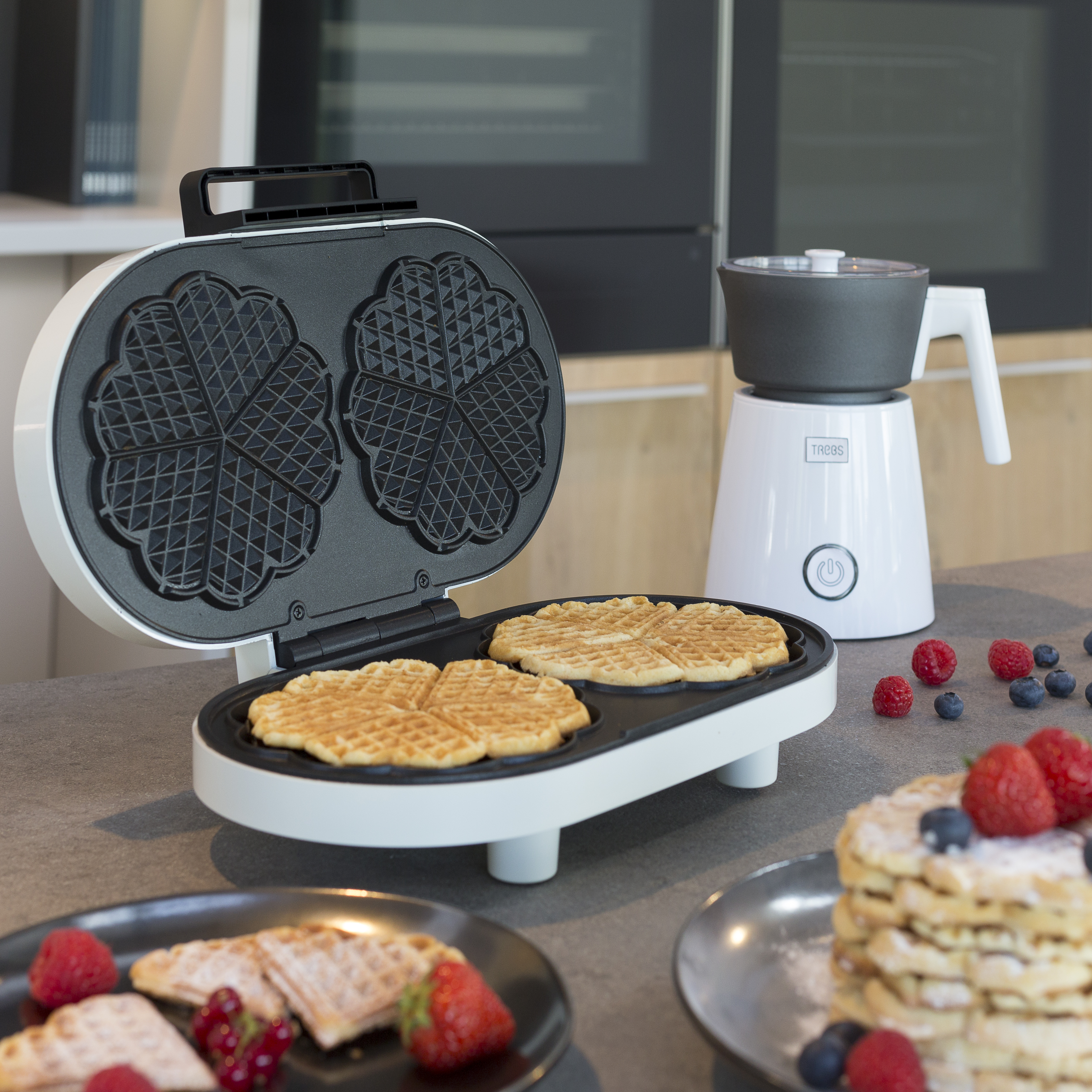 Waffeleisen mit Waffeln darin und ein Milchaufschäumer. Beeren auf dem Tisch.