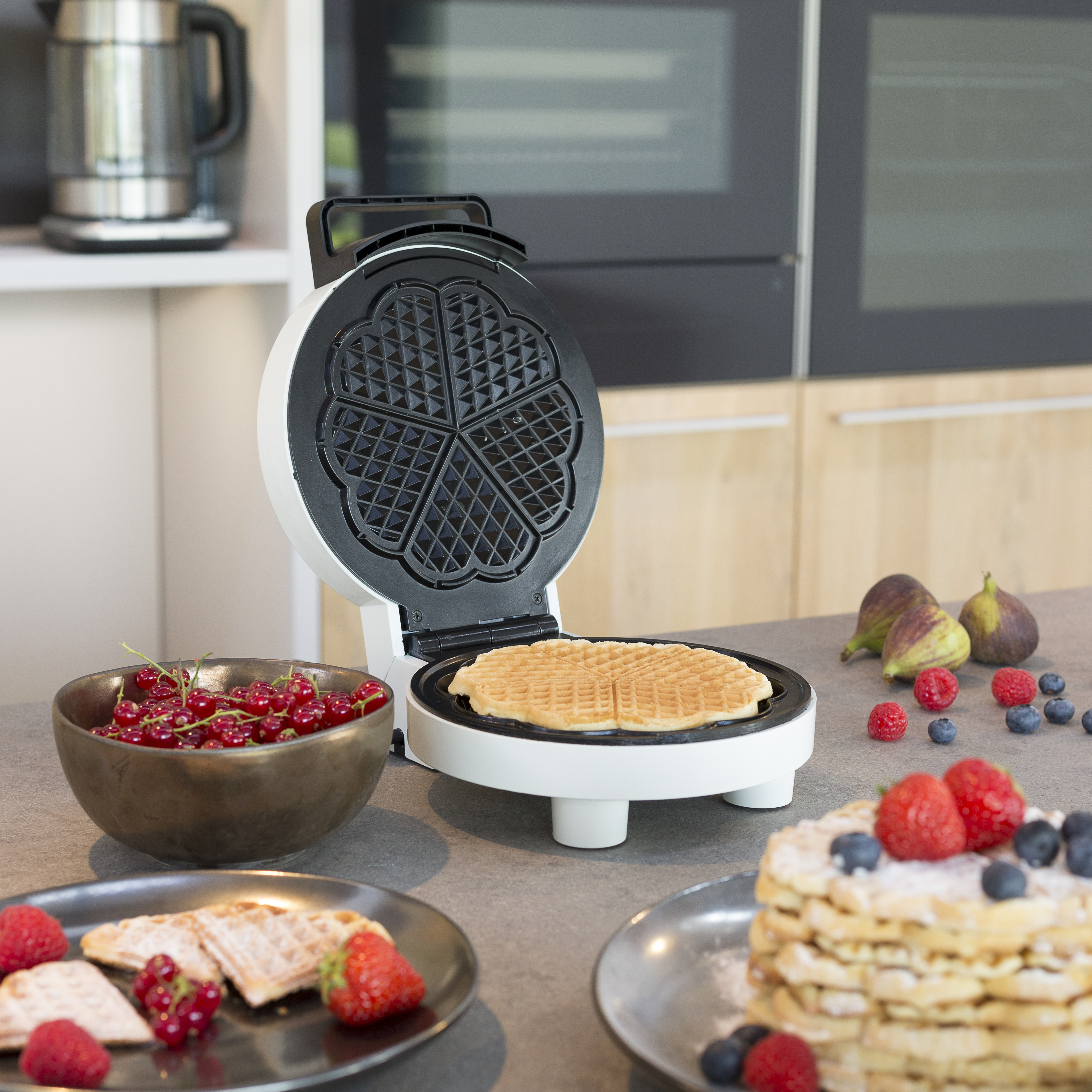 Waffeleisen mit Waffel, Johannisbeeren, Feigen, Himbeeren und Blaubeeren auf grauer Arbeitsplatte.