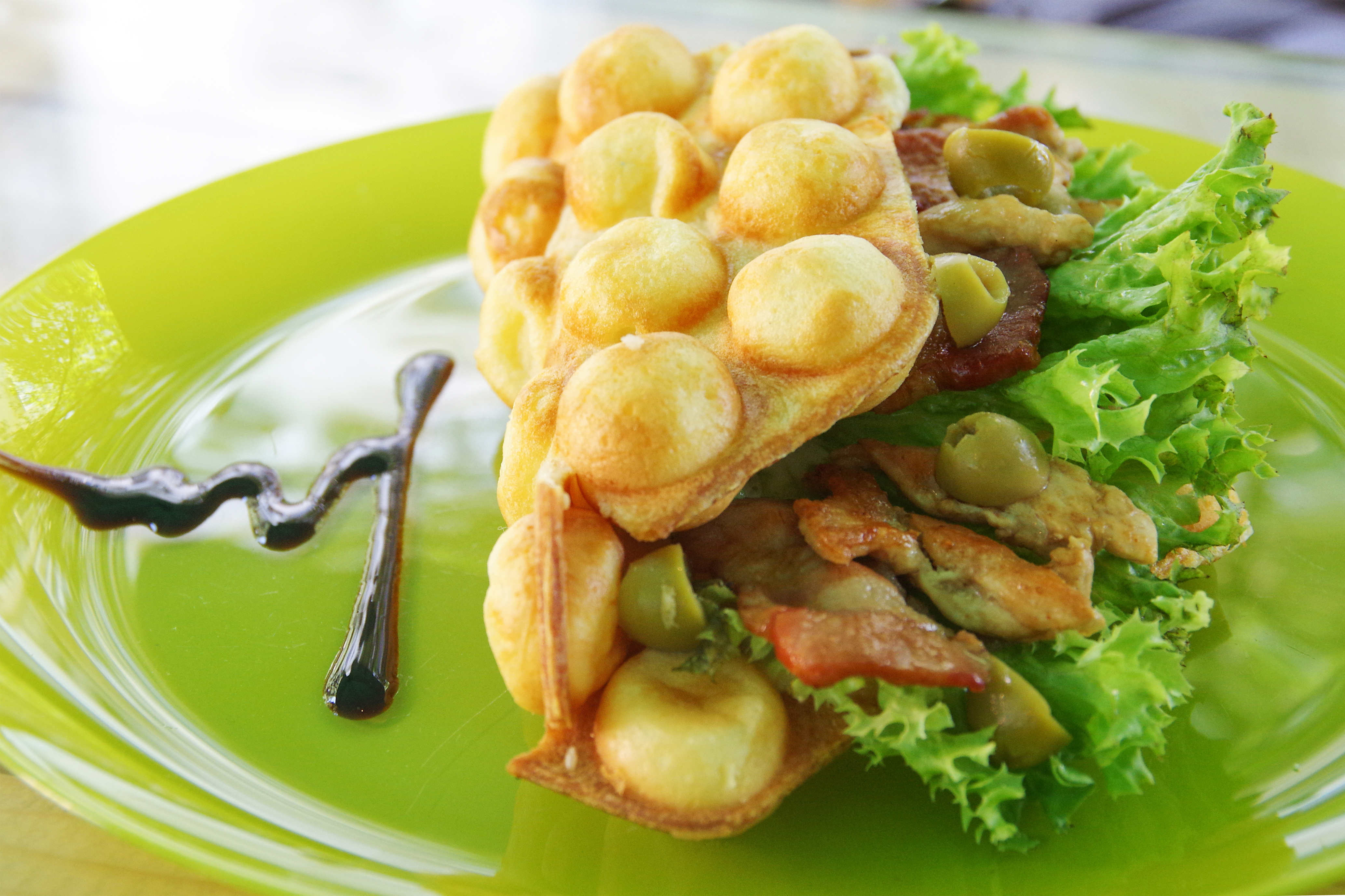 Un plato verde con un gofre relleno de ensalada, aceitunas y carne, con salsa al lado.