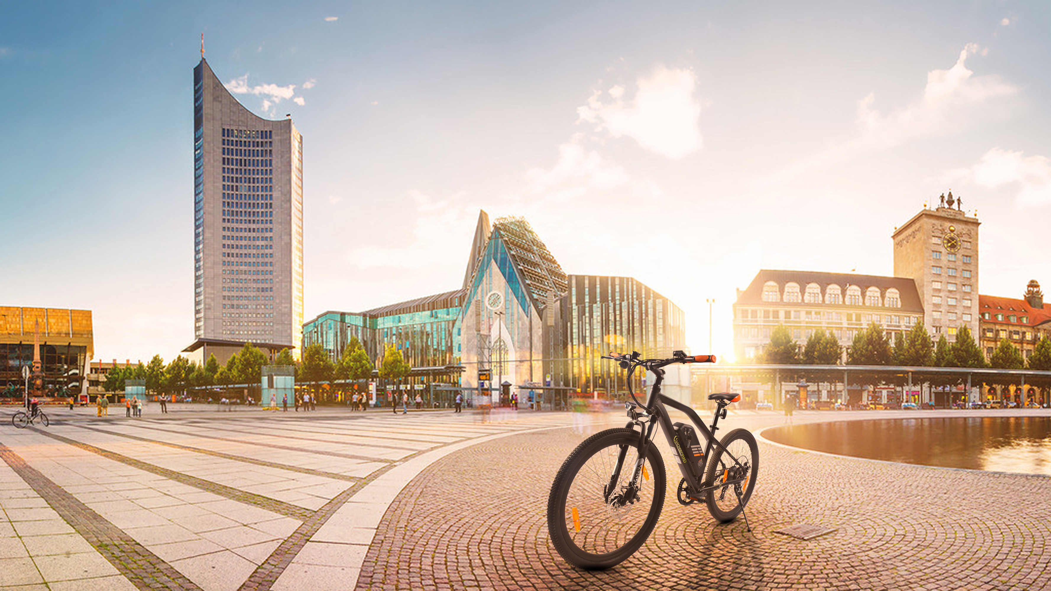 Stadtlandschaft mit modernen Gebäuden, einem Fahrrad und einem weiten offenen Bereich mit Wasserspielen. Blauer Himmel, Sonnenuntergang.