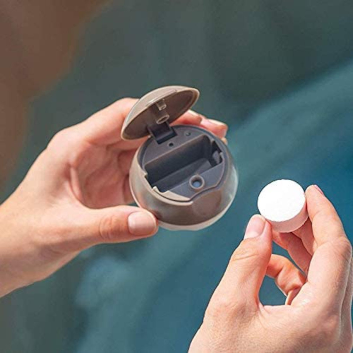 Hände halten einen offenen Behälter und eine weiße Tablette, wahrscheinlich für einen Pool, vor verschwommenem blauem Hintergrund.