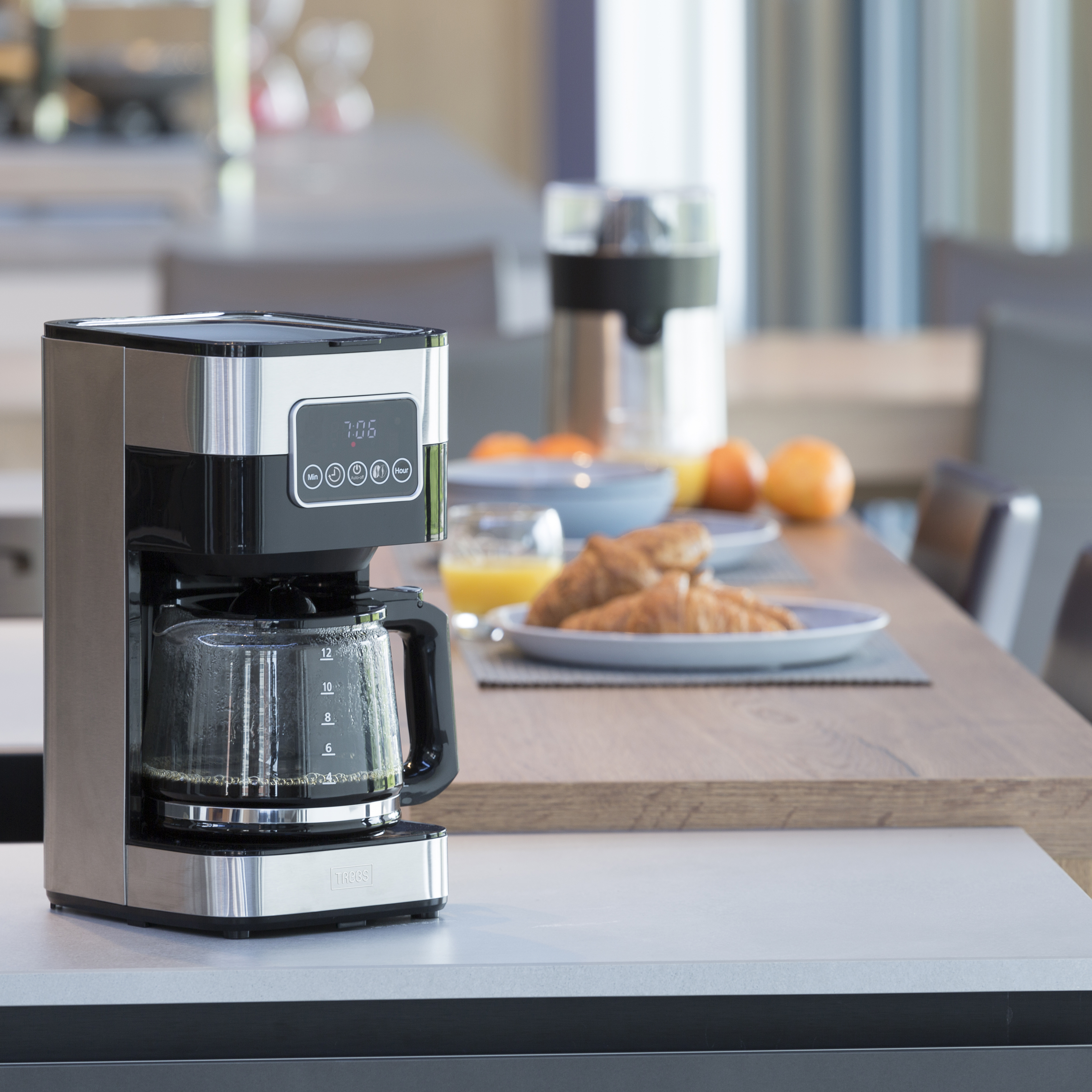Kaffeemaschine auf weißer Oberfläche, Tisch für Frühstück im Hintergrund.