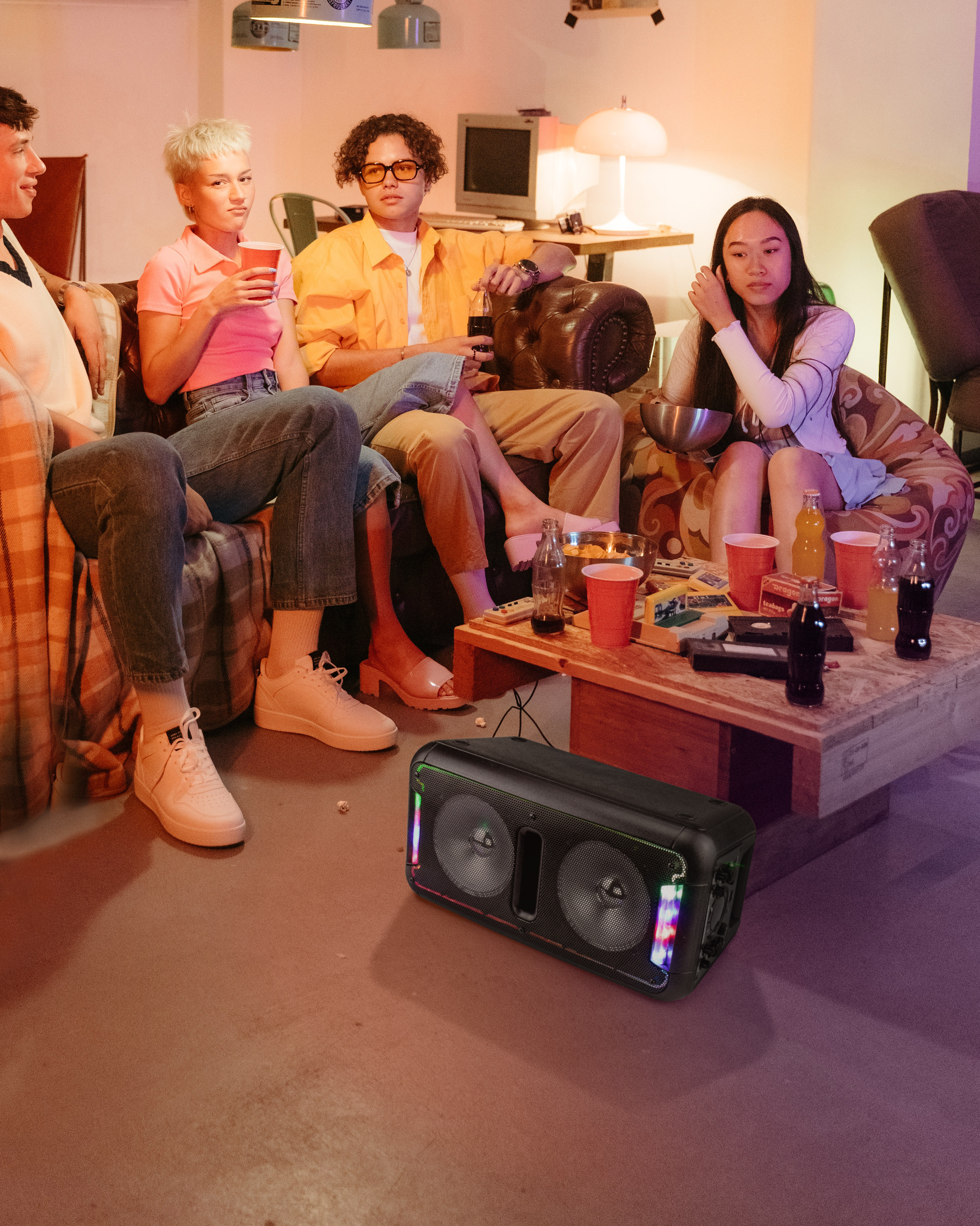 Eine Gruppe von Freunden sitzt um einen Tisch mit Getränken und Snacks, ein Lautsprecher steht auf dem Boden.