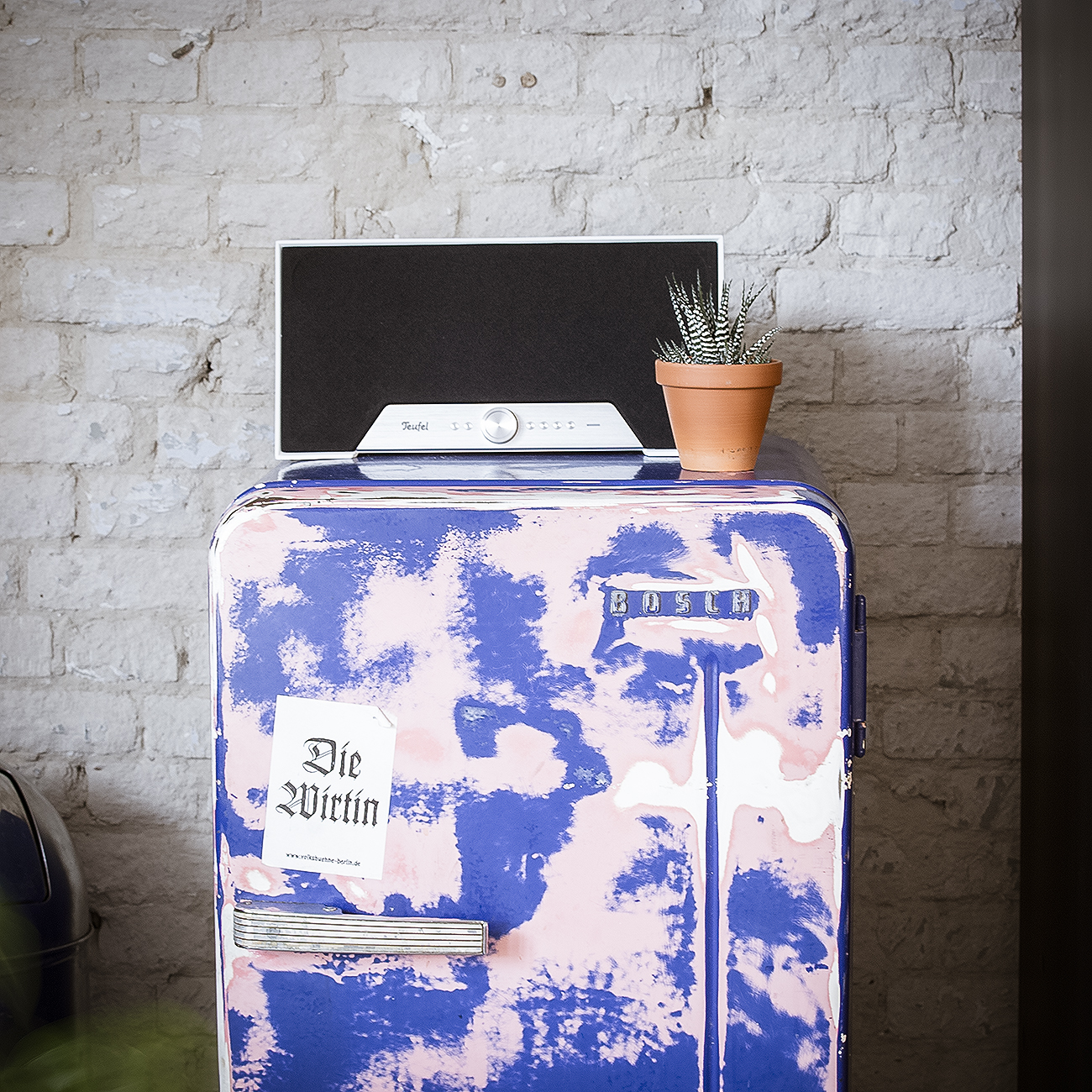 Een luidspreker en een kleine plant op een retro koelkast tegen een bakstenen muur.