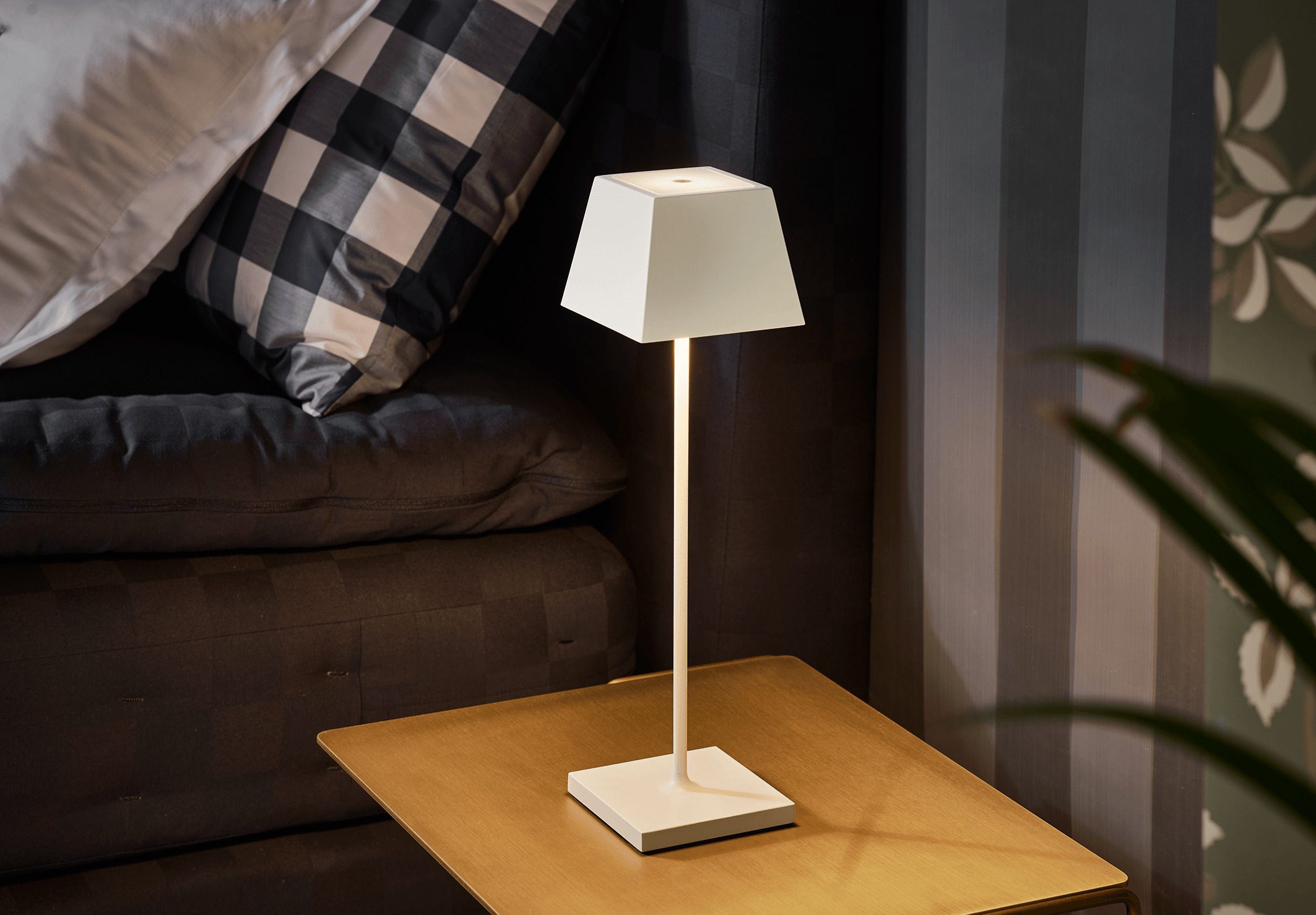 Eine weiße Lampe mit quadratischem Schirm steht auf einem Tisch, in der Nähe eines Bettes mit Kissen.