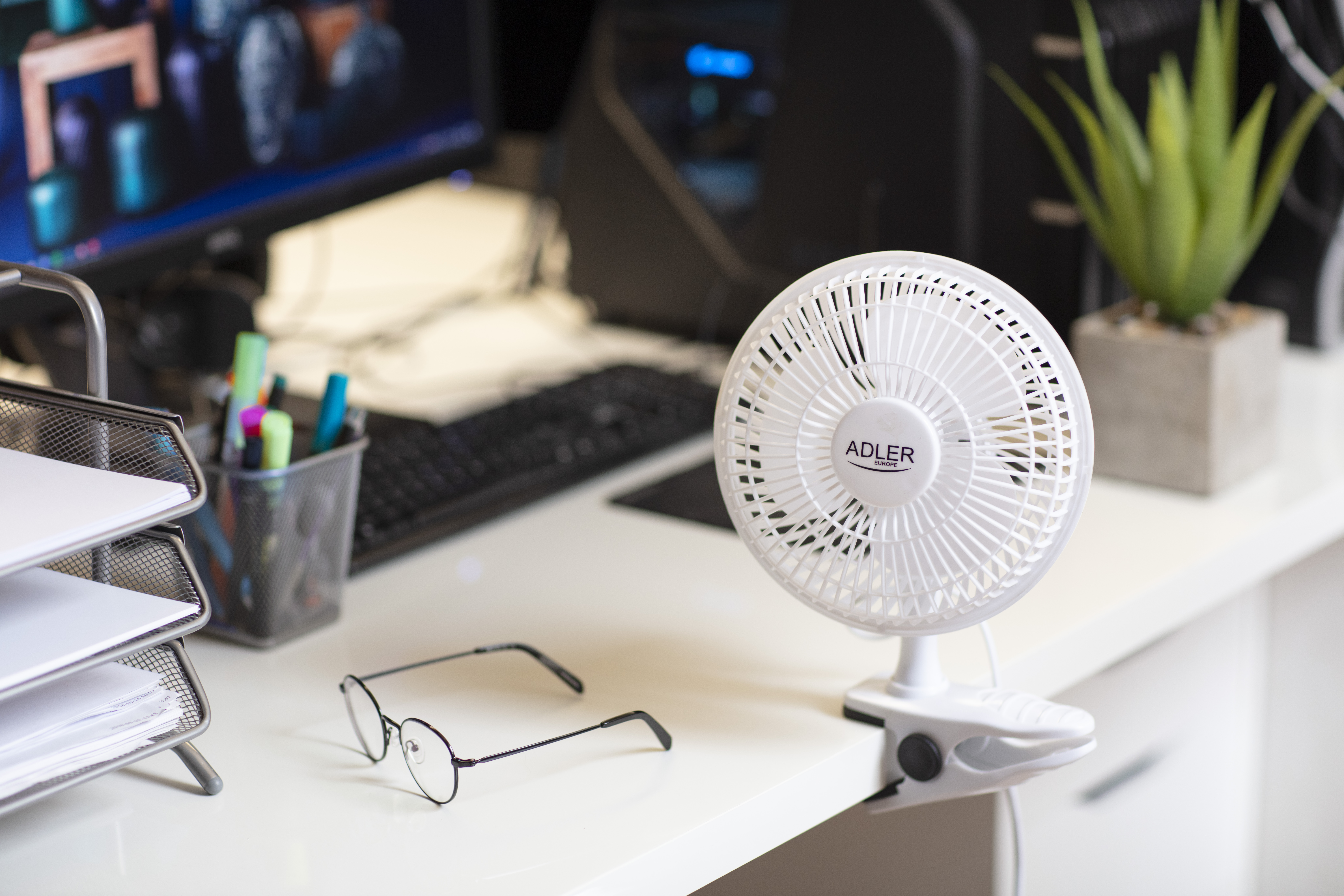 Białe biurko z małym wentylatorem, okularami, monitorem i pojemnikiem na długopisy, tworzące miejsce pracy.