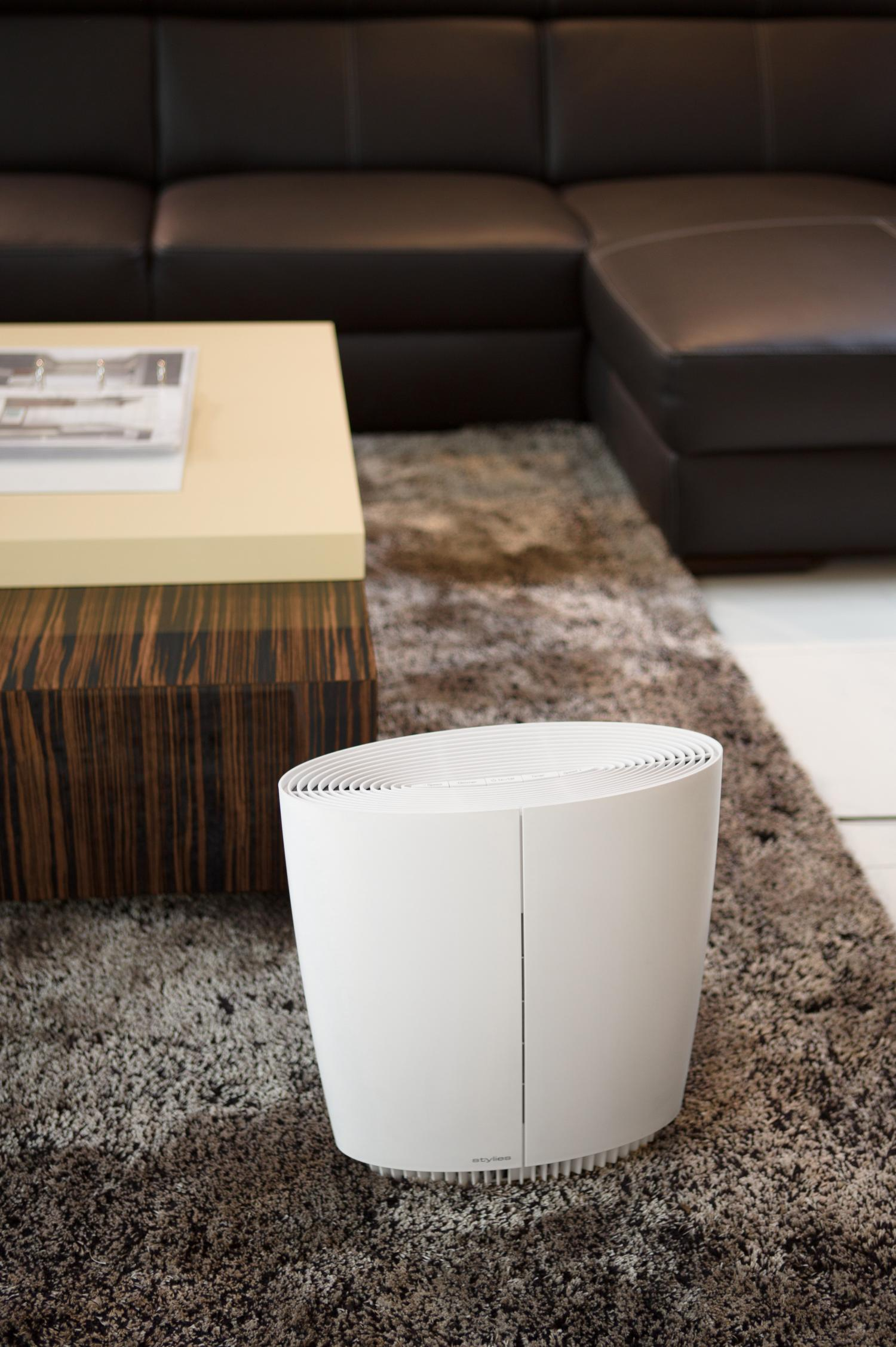 Un purificatore d'aria bianco su un tappeto, con un tavolino e un divano sullo sfondo, creando un ambiente soggiorno.