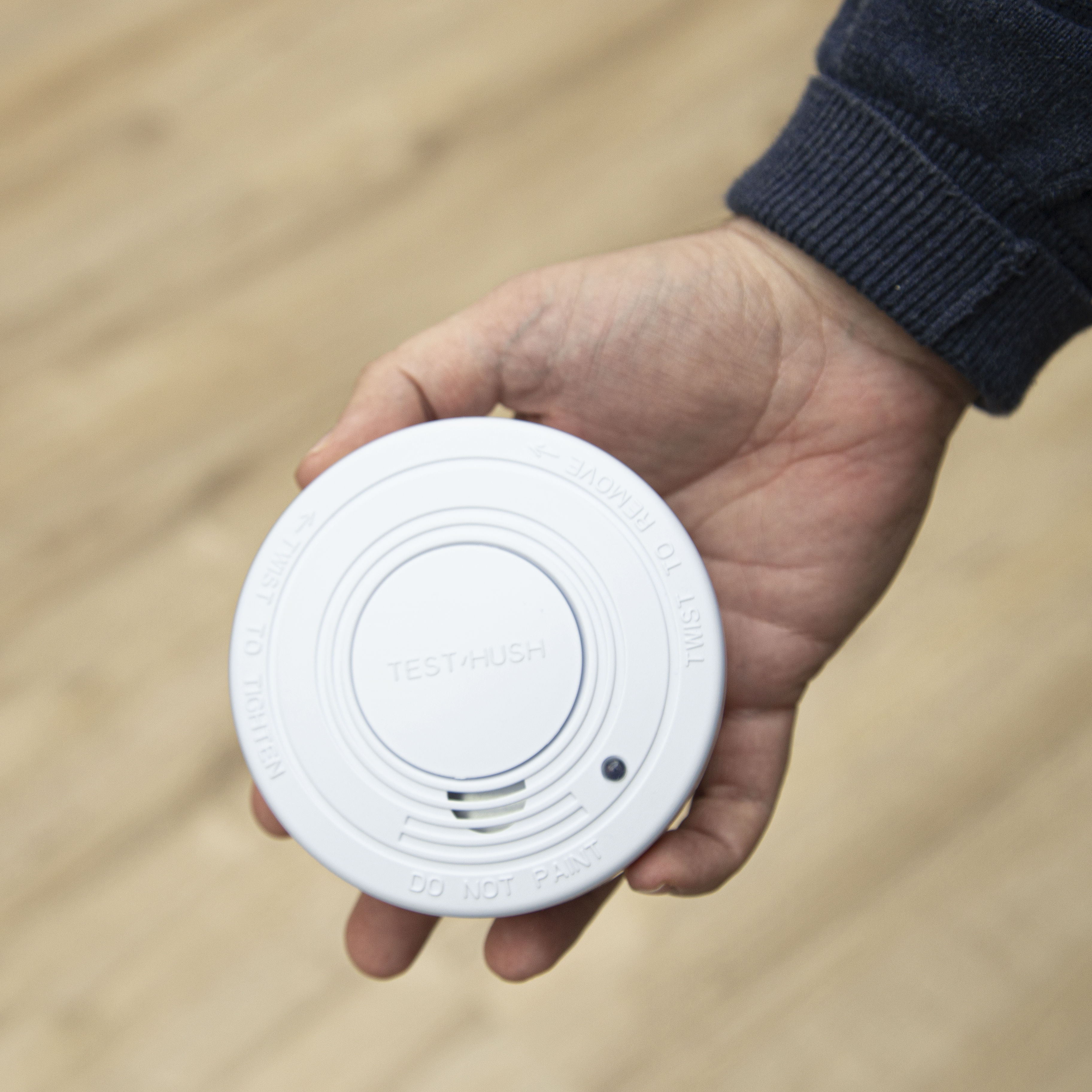 Osoba trzymająca białą czujkę dymu z napisem 'TEST+HUSH' i 'DO NOT PAINT'.