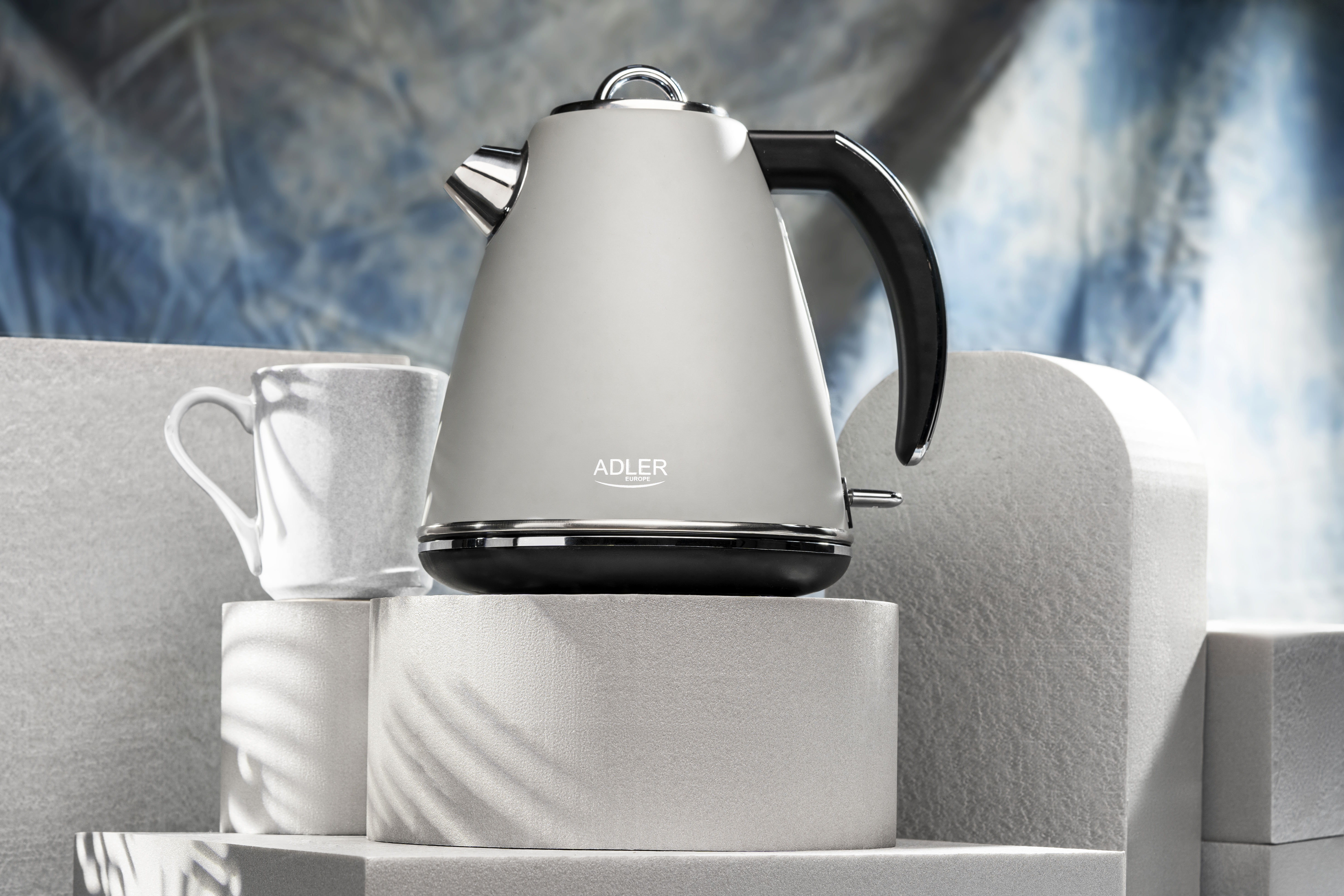 Ein weißer und silberner Wasserkocher steht auf einer weißen Oberfläche mit einer Kaffeetasse.