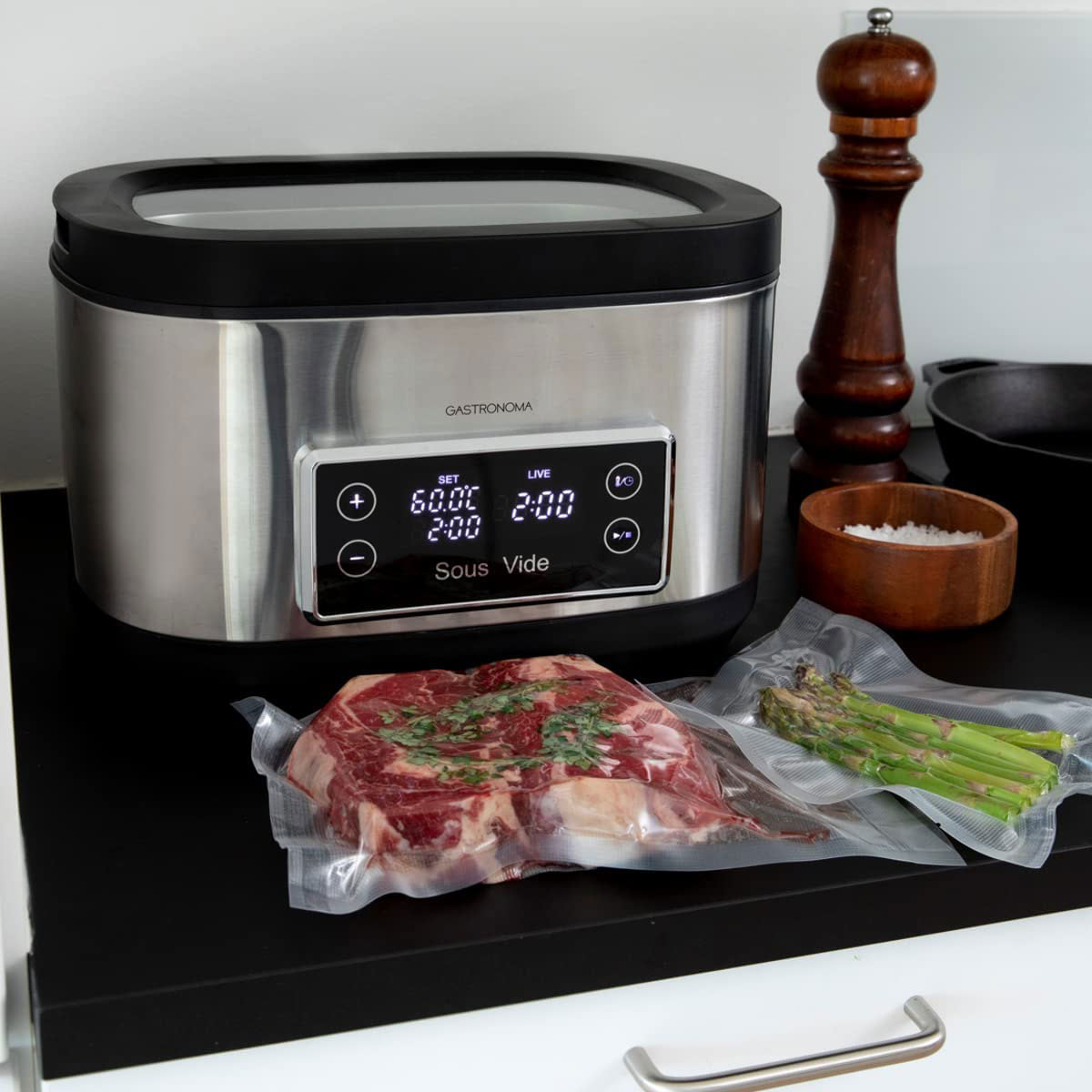 Sous vide machine with steak and asparagus in bags. Salt and pepper are also on the counter.