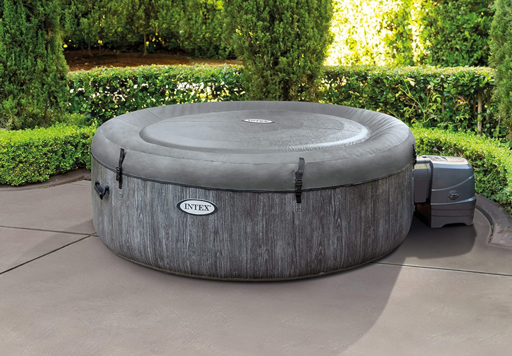 Ein Whirlpool im Freien mit holzartiger Außenseite und grauer Abdeckung. Es befindet sich auf einer Terrasse mit Grün.