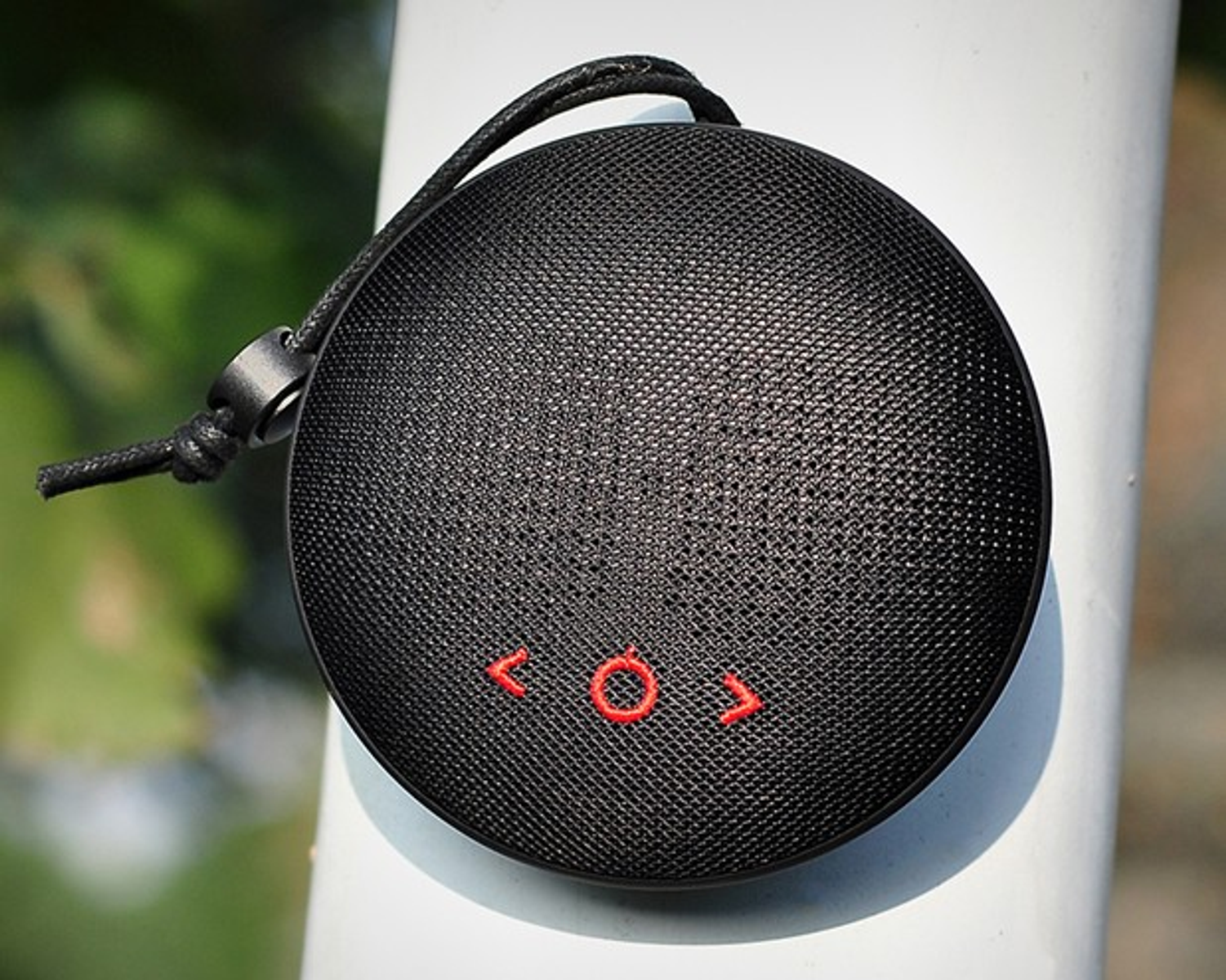 Un altavoz negro redondo con botones rojos cuelga de un poste blanco. Tiene una correa negra.