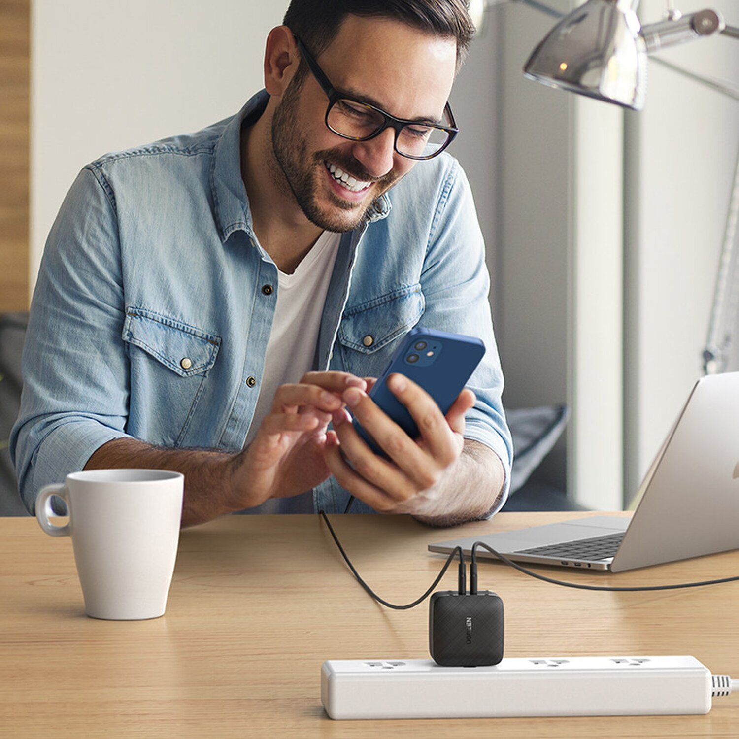 Ein lächelnder Mann, der ein Smartphone benutzt. Ein Laptop, eine Steckdosenleiste und ein Becher auf dem Tisch.