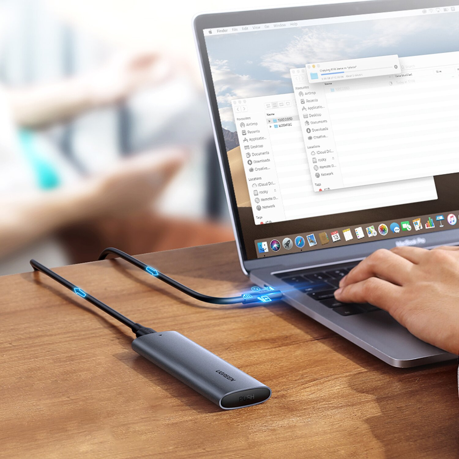 Laptop, verbunden mit grauem UGREEN-Gerät über blau leuchtendes Kabel. Hand am Keyboard.