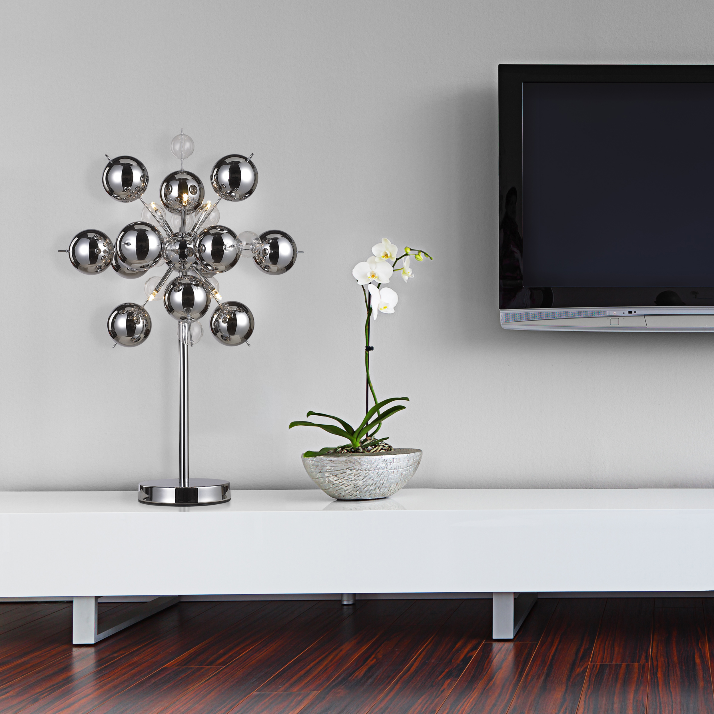 Eine Chromlampe in einem Raum mit weißem Schrank, TV und Pflanze. Dahinter eine weiße Wand.