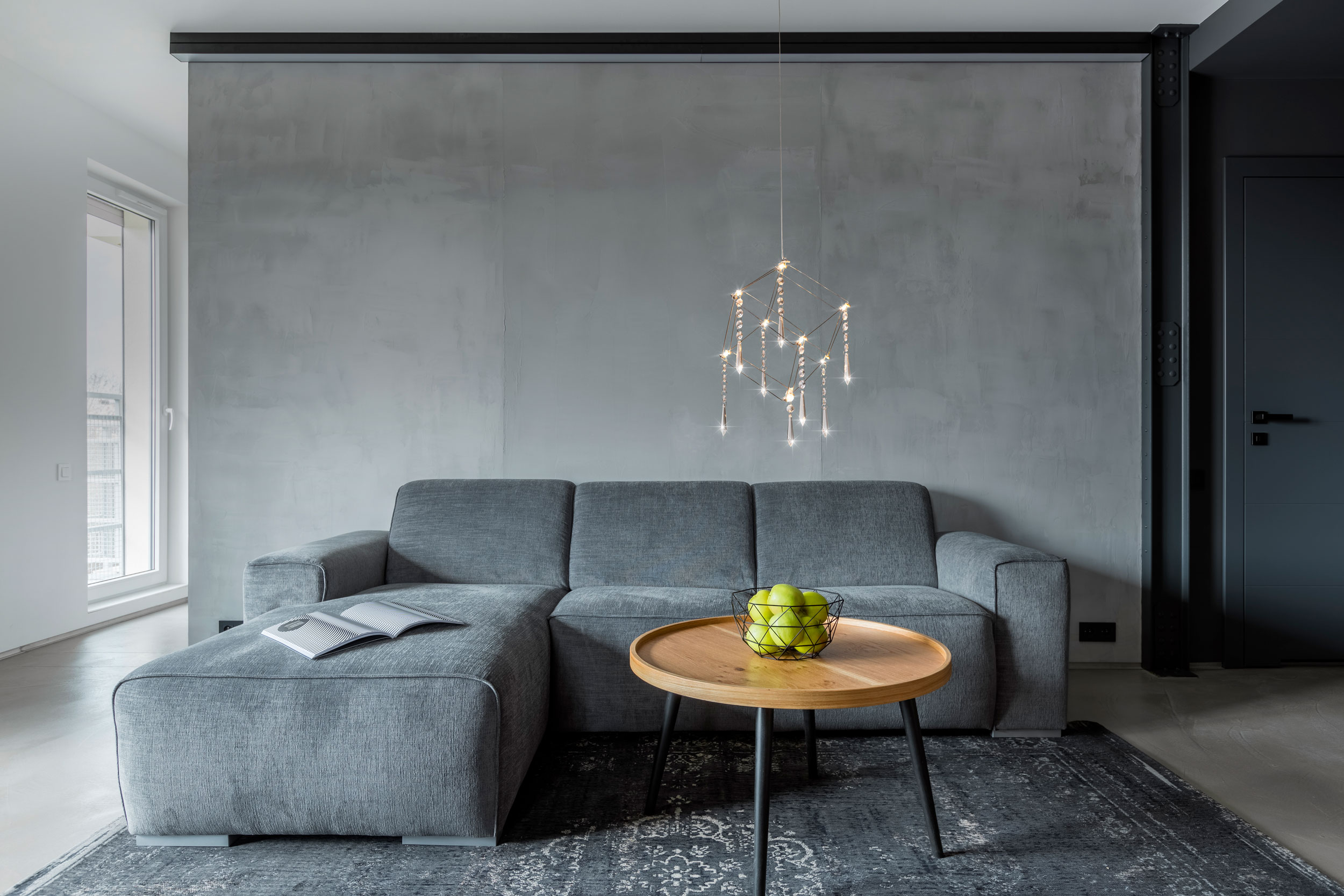 Graues Ecksofa, runder Holztisch und grüne Äpfel. Graue Wand im Hintergrund.