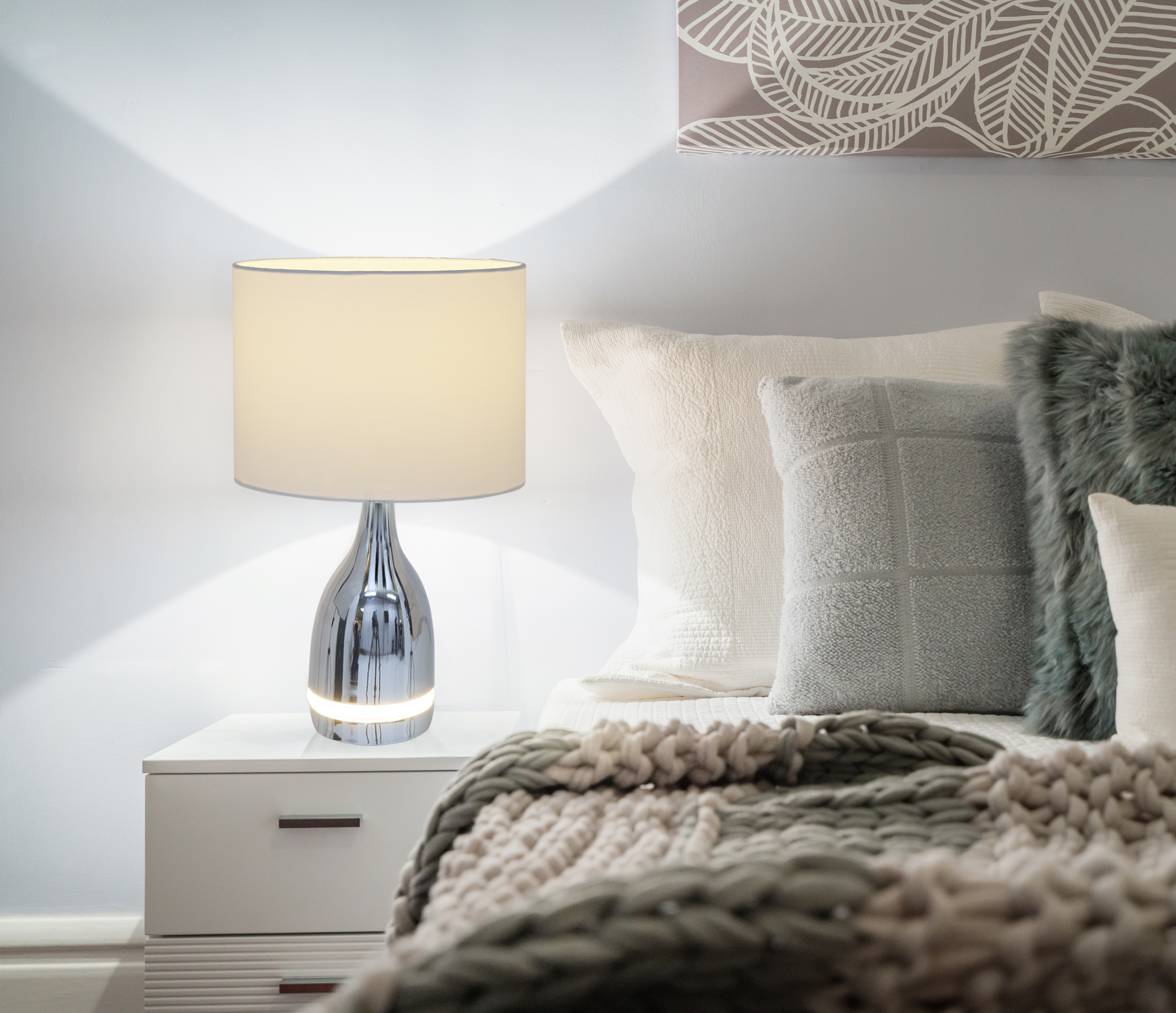 Eine Lampe auf einem Nachttisch in einem Schlafzimmer. Die Lampe hat einen weißen Schirm und einen Chromfuß.