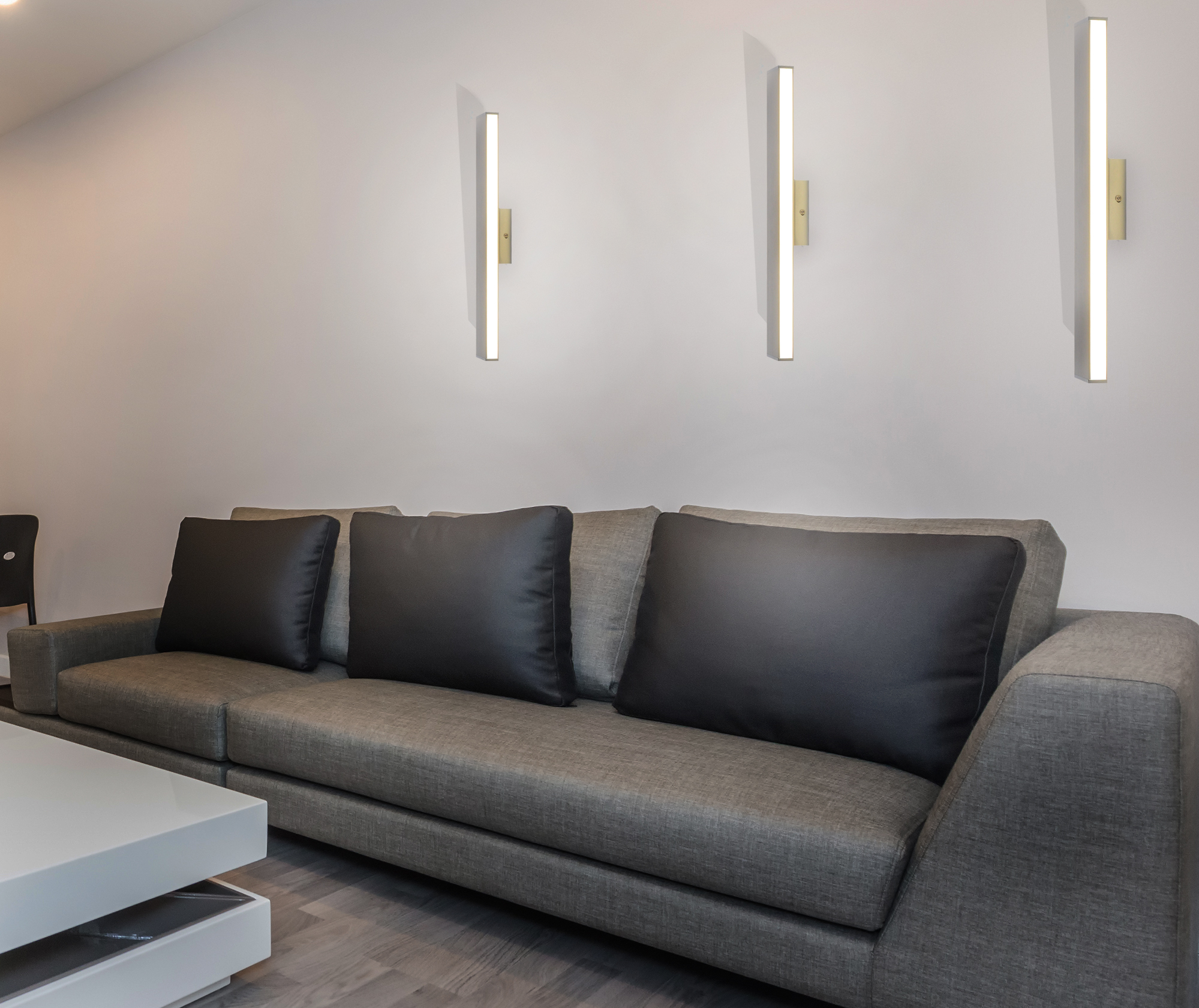 Graues Sofa mit schwarzen Kissen unter drei Wandleuchten an einer weißen Wand.