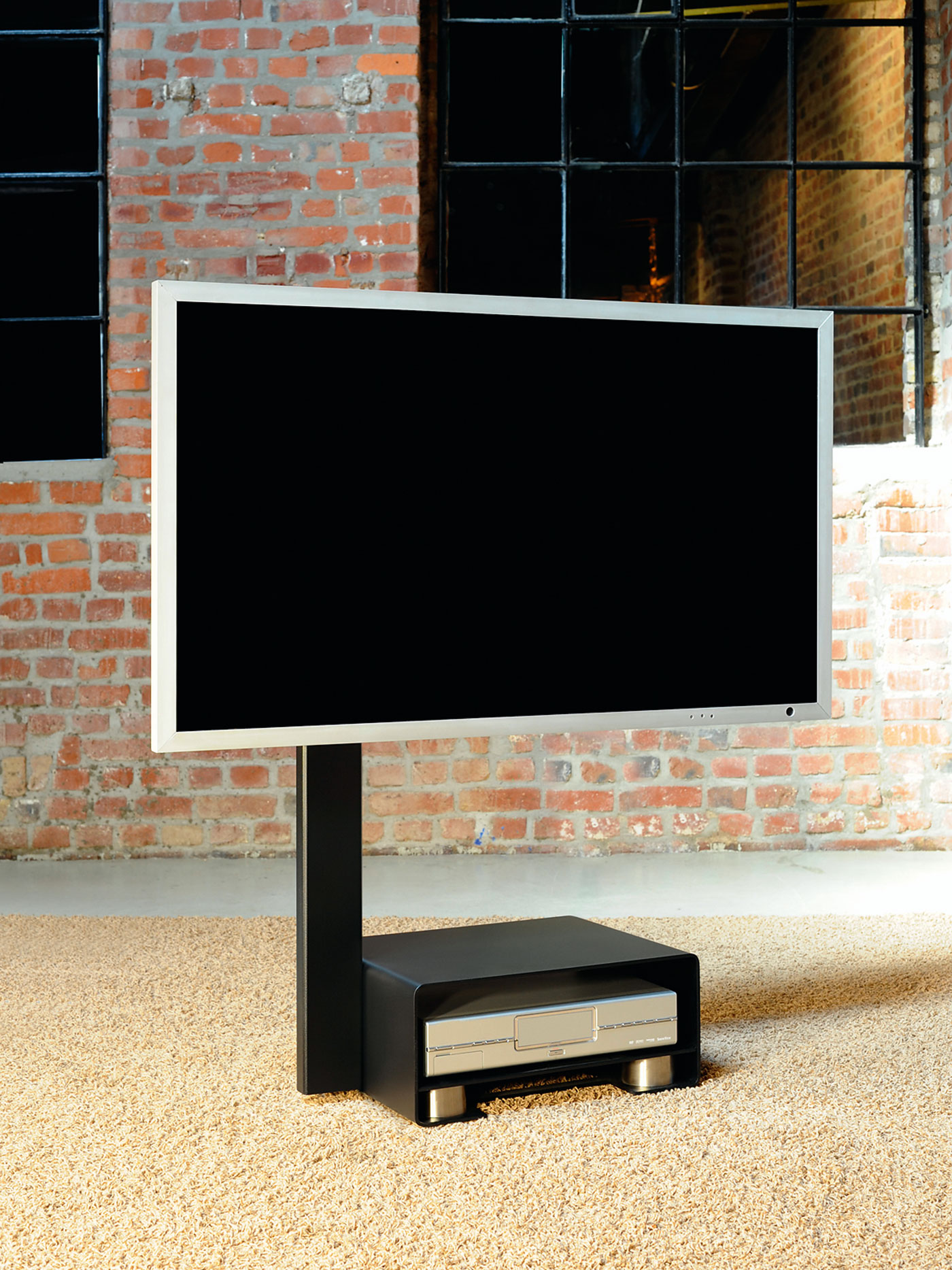 Ein Fernseher auf einem schwarzen Ständer vor einer roten Ziegelmauer. Der Boden ist mit Teppich bedeckt.