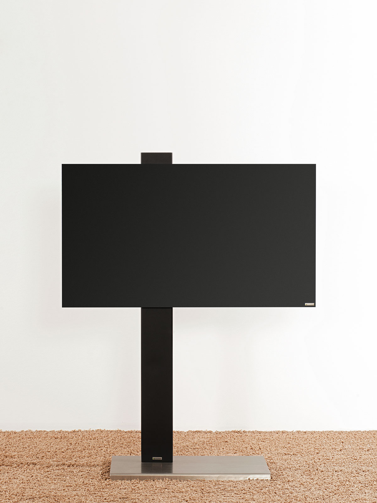 Schwarzer Fernseher auf einem Ständer, auf beigem Teppich vor weißer Wand.
