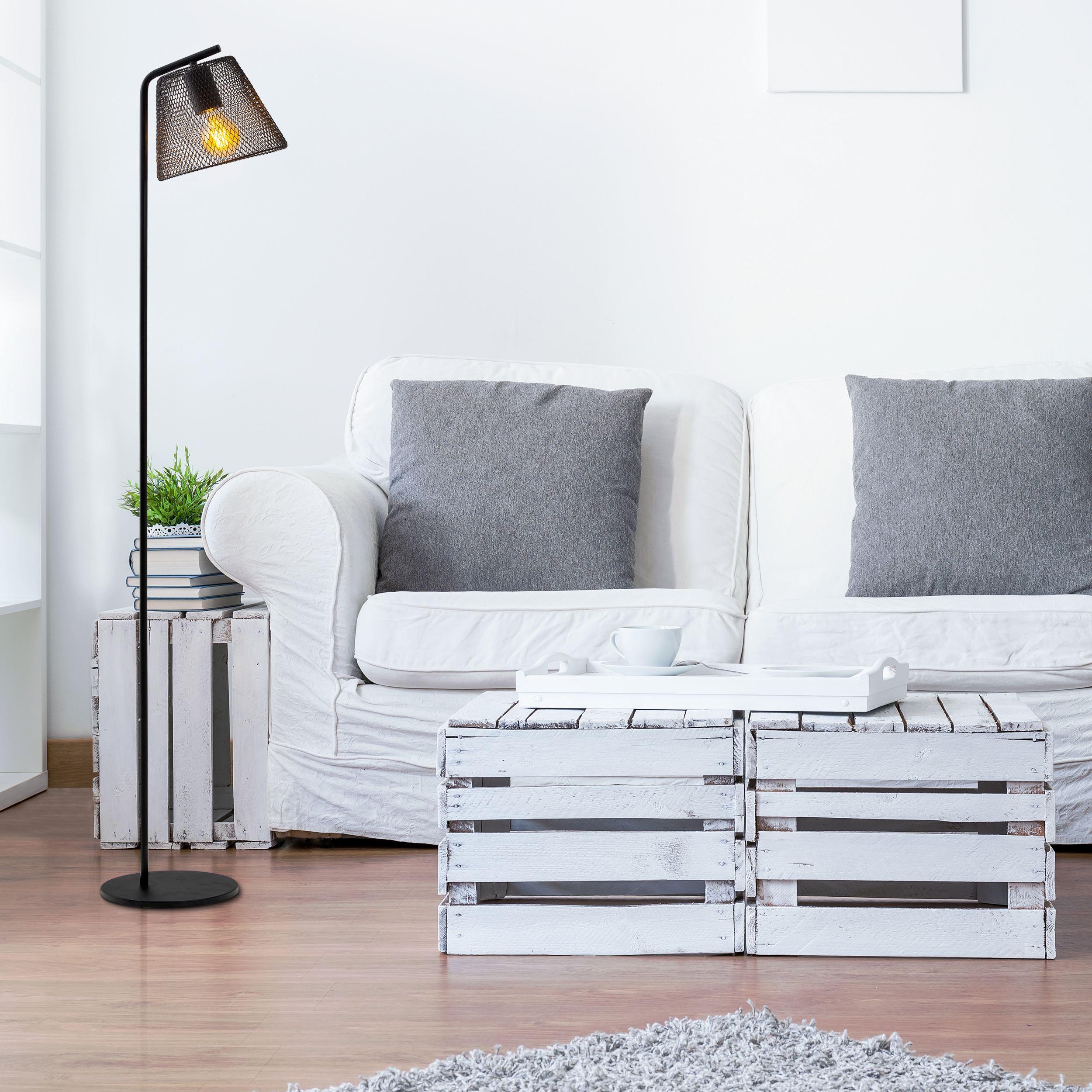 Eine Stehlampe mit einem Lampenschirm aus Netzstoff. Ein weißes Sofa, graue Kissen und ein Tisch aus Holzkisten sind im Bild.