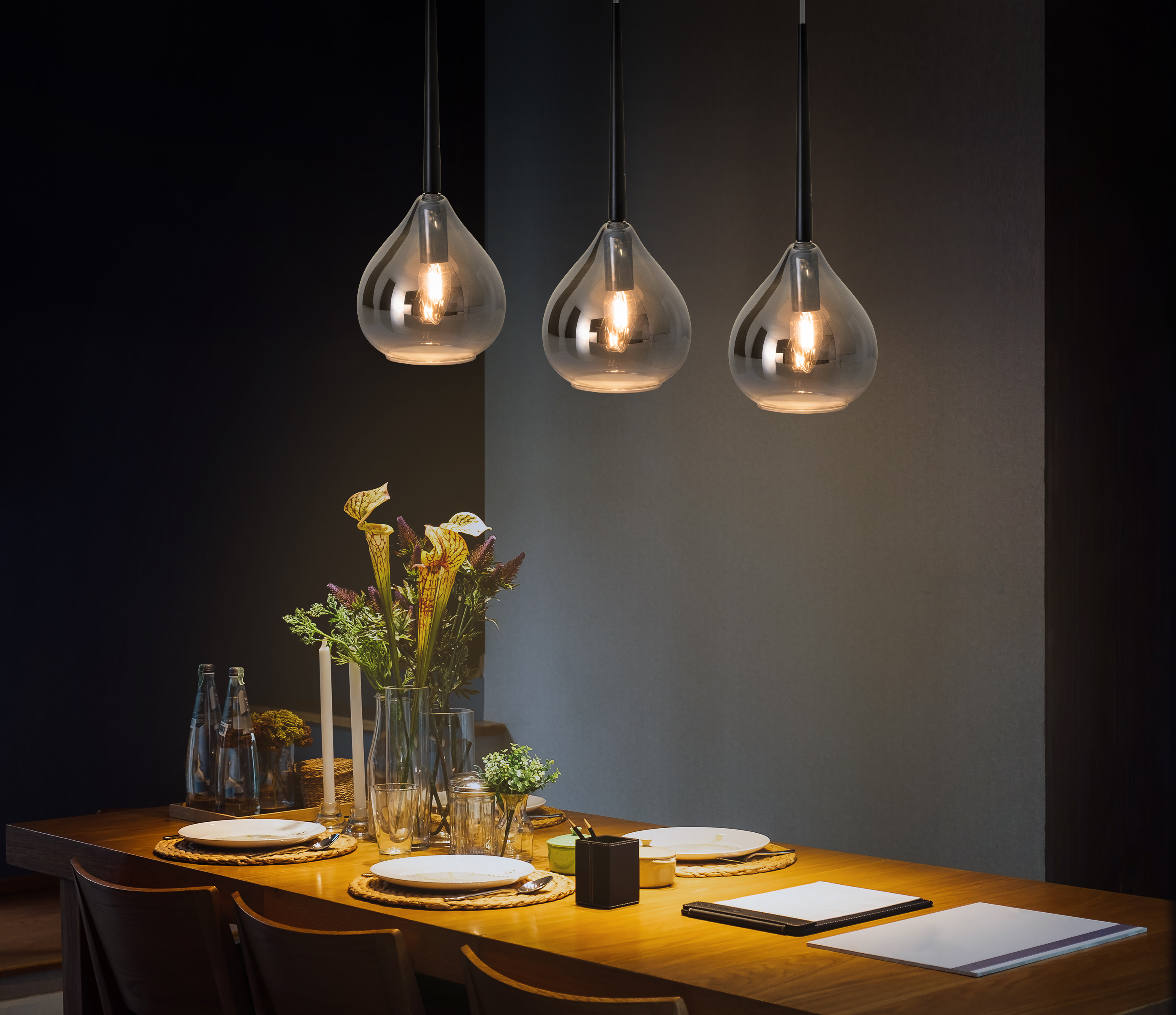 Trois lampes suspendues au-dessus d'une table en bois dressée pour le dîner, avec des fleurs et des bougies.