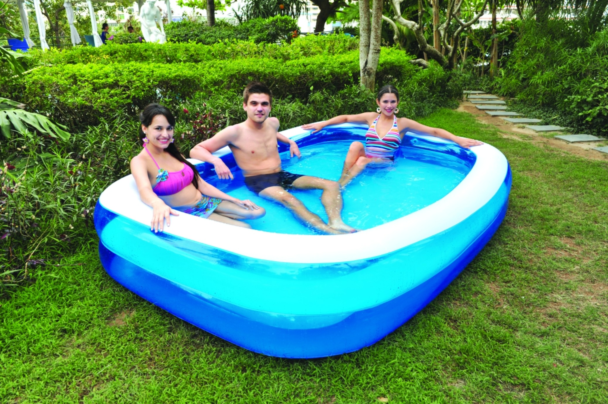 Drei Personen in einem blau-weiß herzförmigen aufblasbaren Pool, in einer Gartenumgebung.
