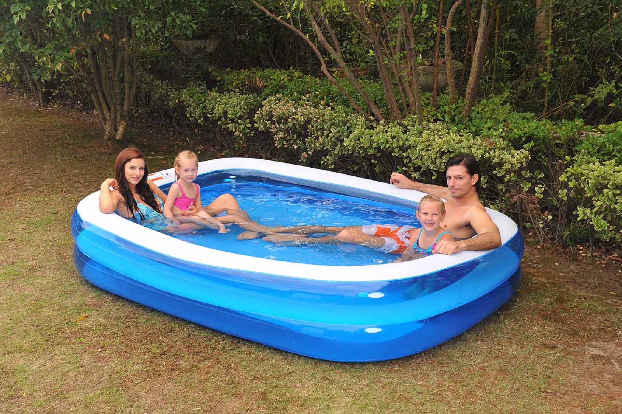 Familie in einem blau-weißen rechteckigen Pool, umgeben von Grün, genießt das Wasser.