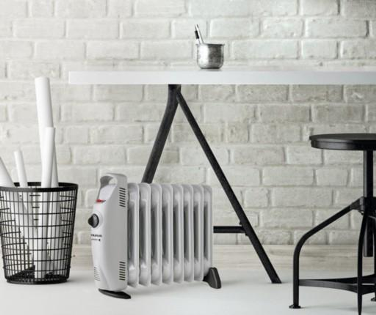 Biały grzejnik olejowy przed ceglaną ścianą, obok biurka i stołka.