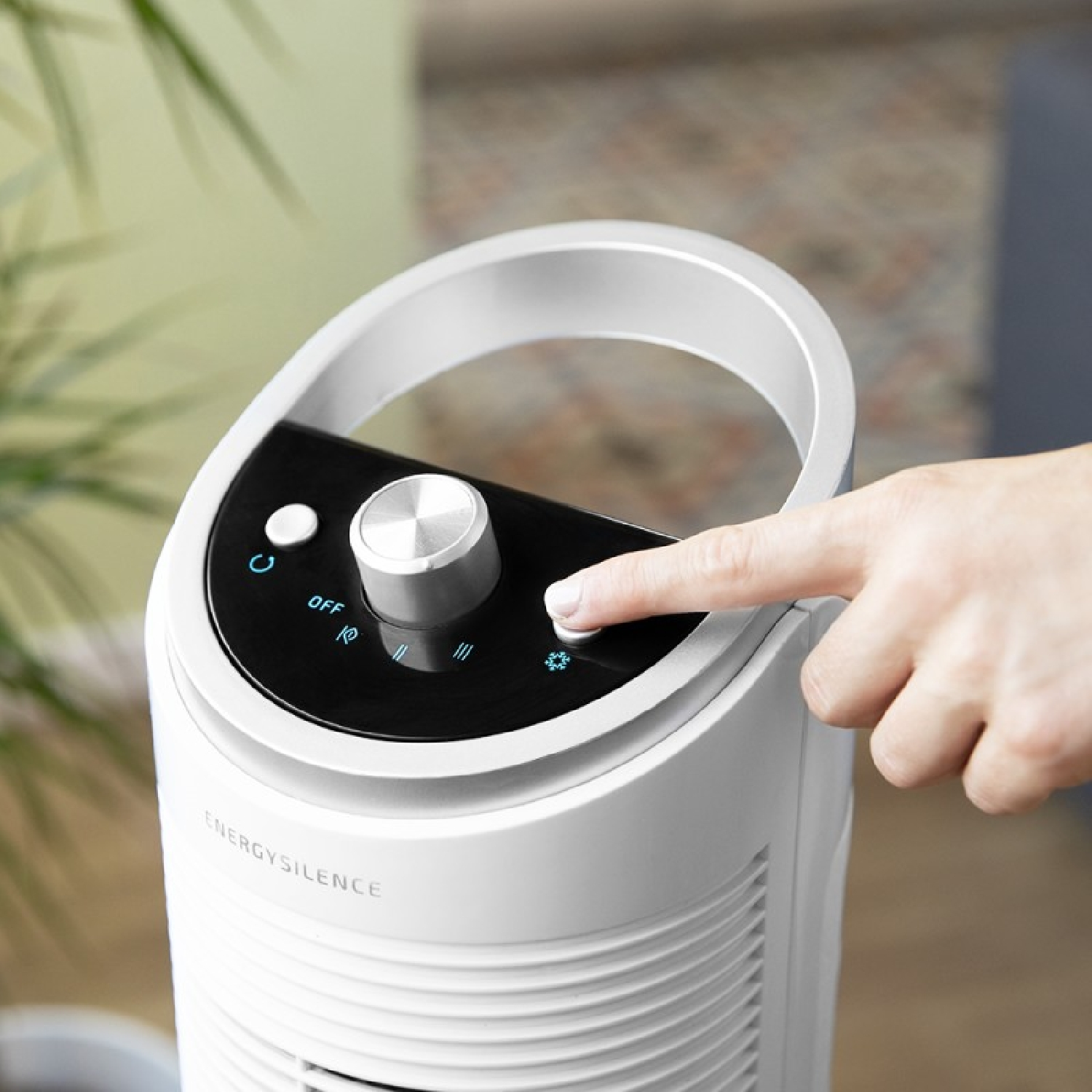 Nahaufnahme eines weißen Ventilators mit silbernem Knopf. Ein Finger drückt eine Taste auf dem Bedienfeld, vor hellem Hintergrund.