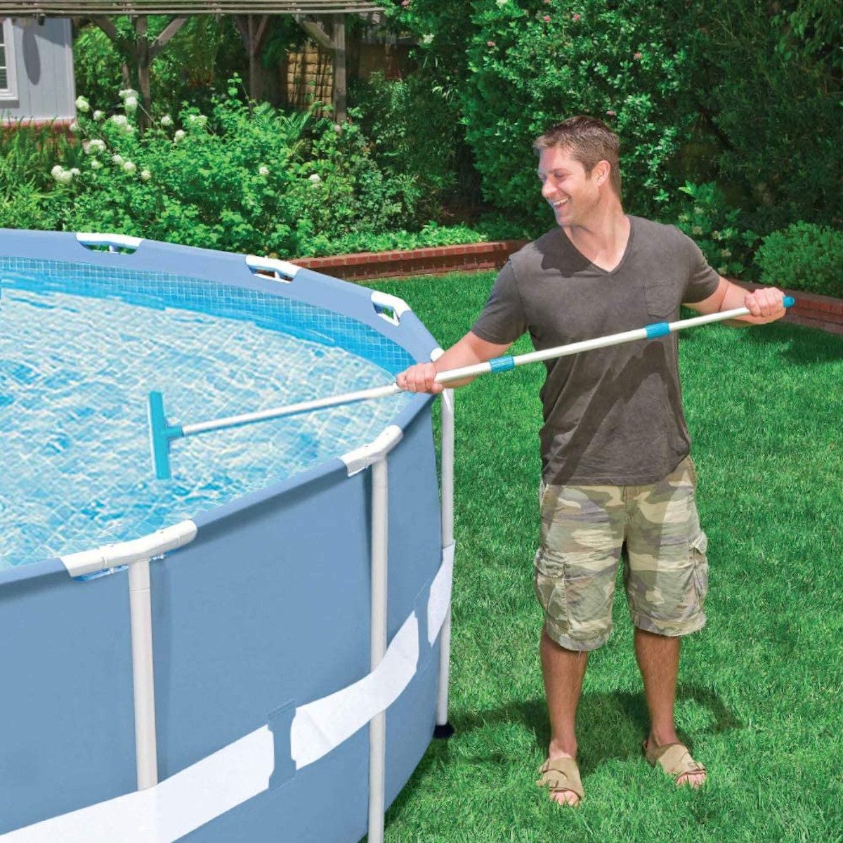 Uomo che pulisce una piscina fuori terra blu con uno skimmer a manico lungo, sfondo di erba verde.