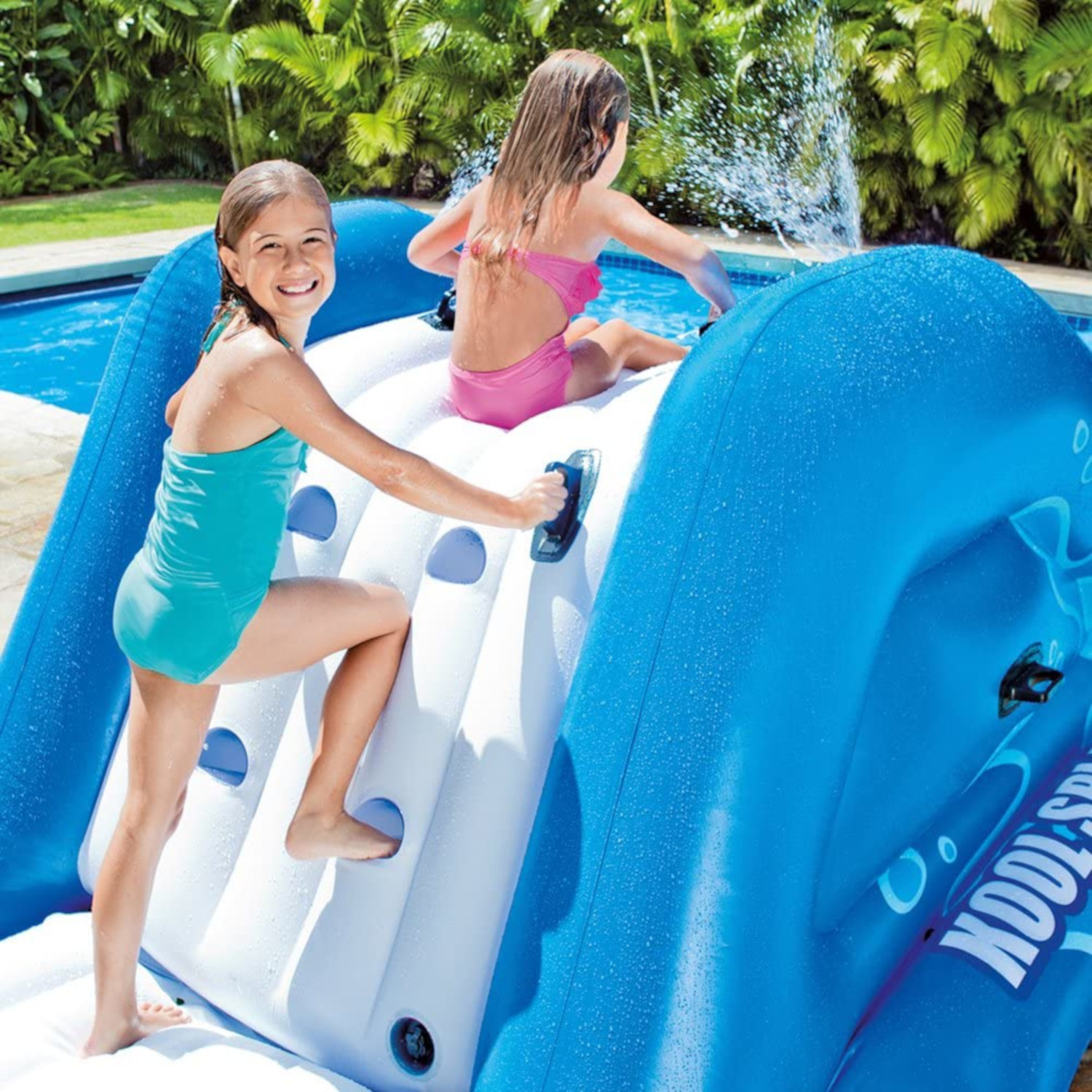 Zwei Mädchen auf einer blauen aufblasbaren Wasserrutsche am Pool. Eins klettert, das andere rutscht. Sonniger Tag.