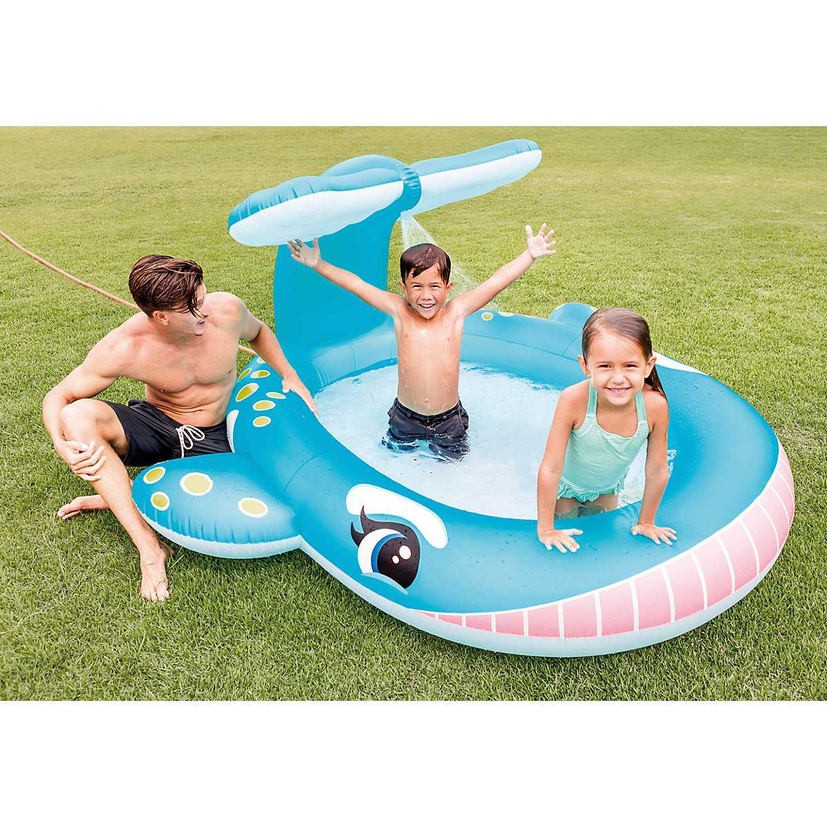 Kinder spielen in einem walförmigen Pool. Ein Mann sitzt in der Nähe des Pools. Grünes Gras im Hintergrund.