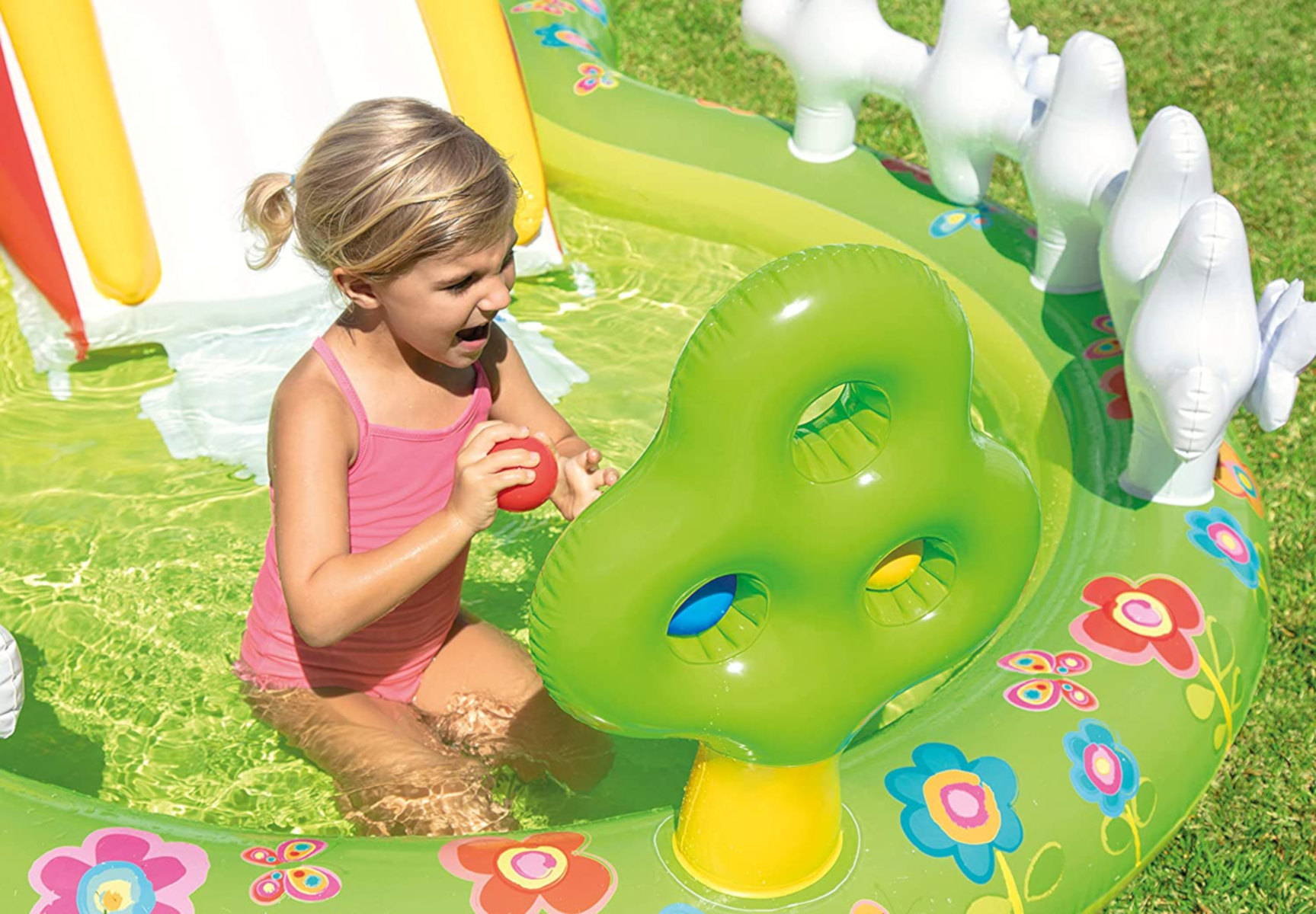 Una bambina in costume da bagno rosa gioca in una colorata piscina gonfiabile con un albero e delle anatre.
