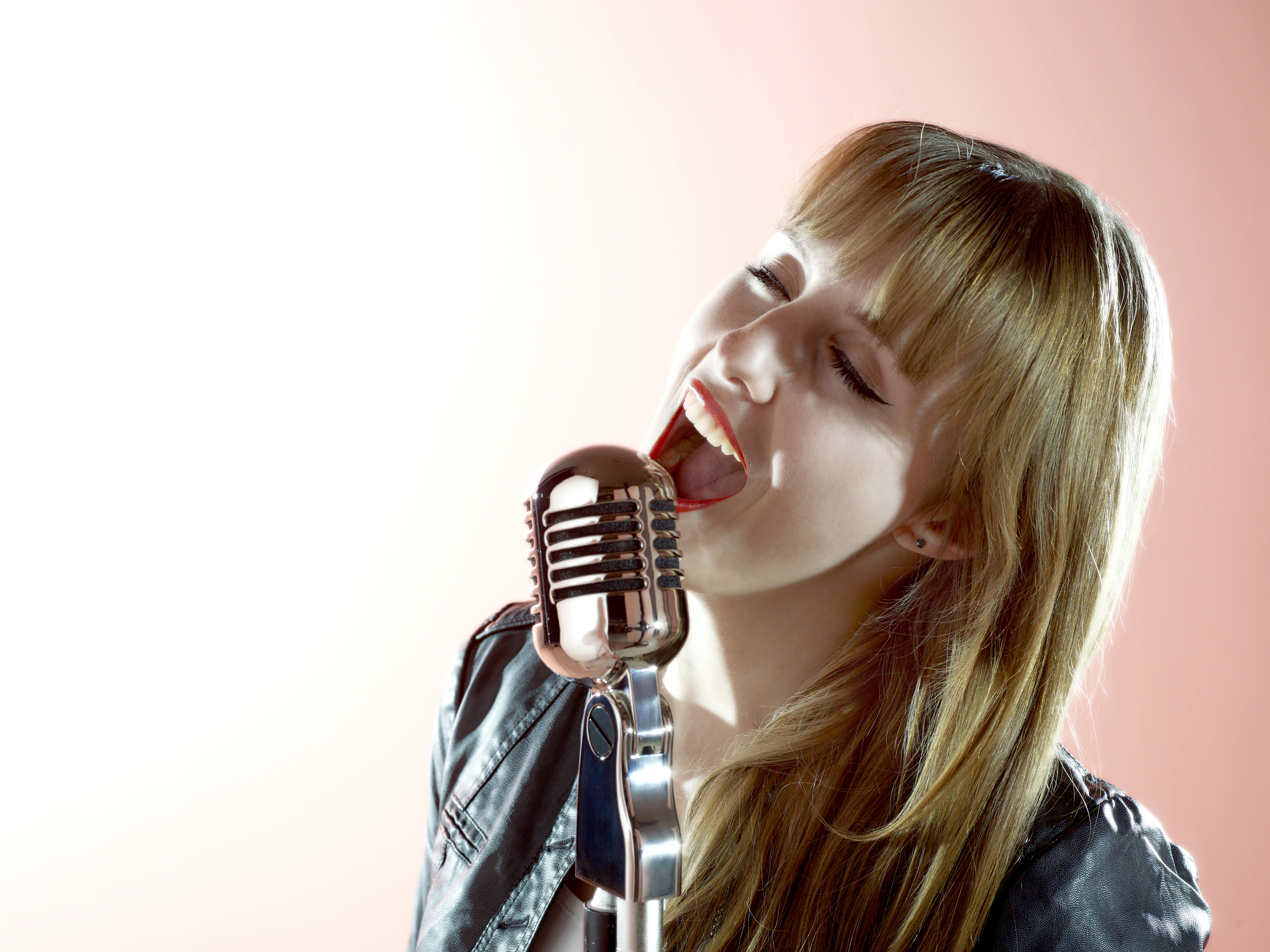 Een vrouw zingt in een zilveren vintage microfoon, mond open, draagt een leren jas.