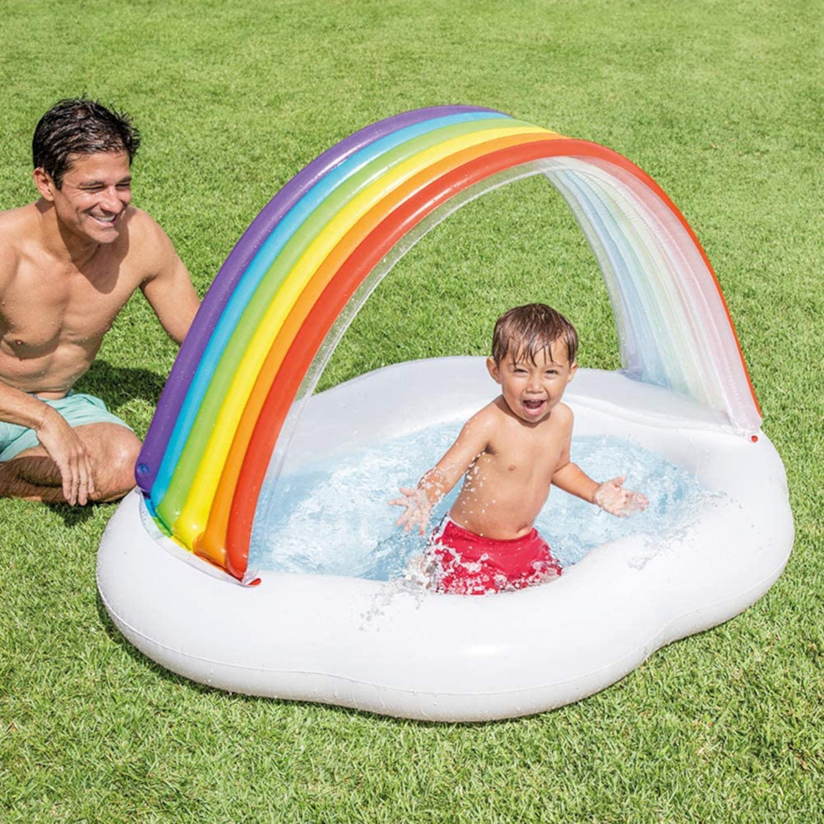 Ein Junge in roten Shorts spielt in einem weißen Planschbecken mit Regenbogenbogen.