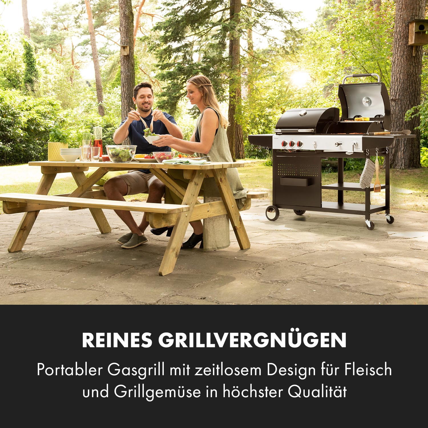 Ein Paar am Picknicktisch mit Essen, Grill und Bäumen im Hintergrund.