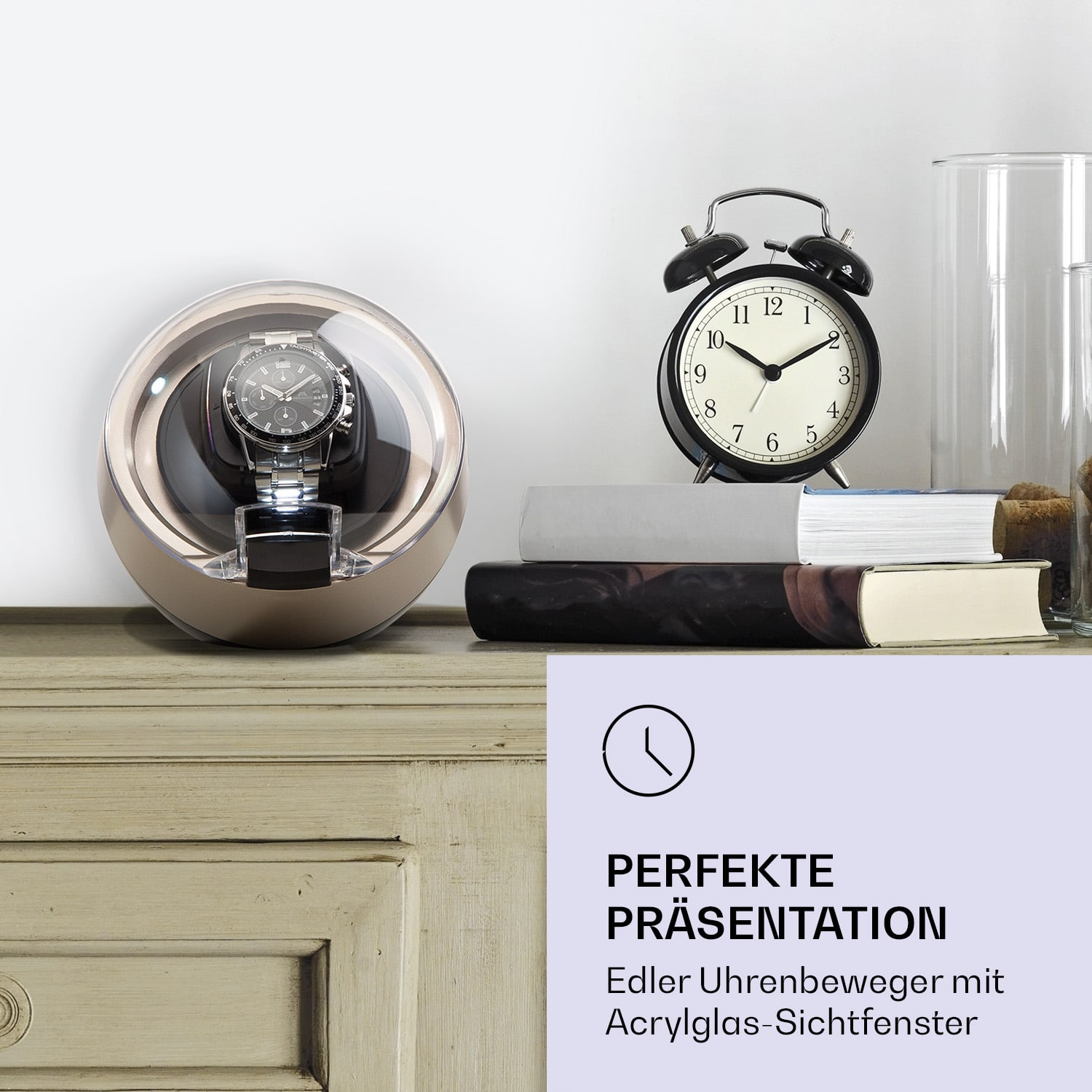 Eine Uhr wird in einem rotierenden Gehäuse mit einem schwarzen Wecker und Büchern auf einem Tisch angezeigt.