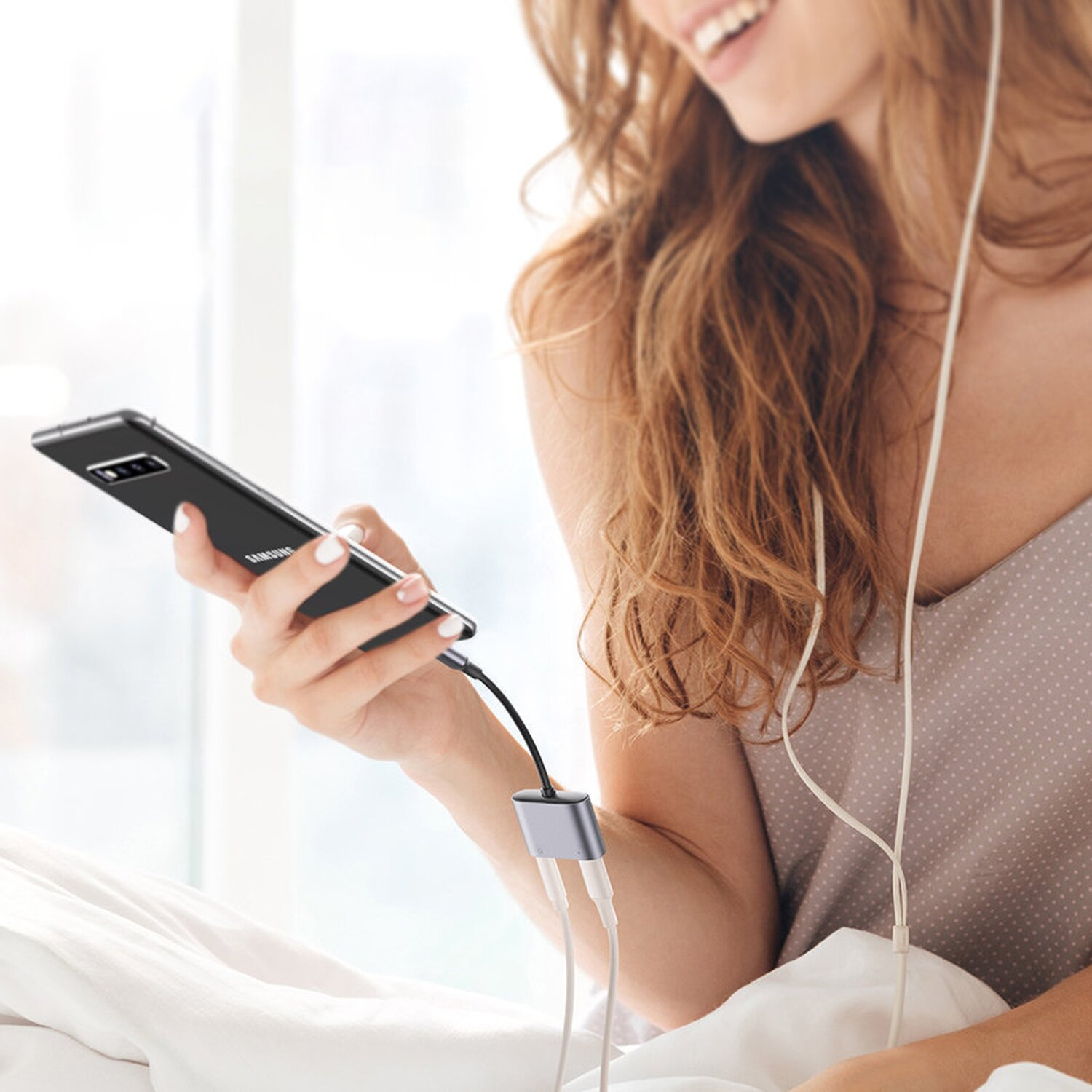 Kobieta trzyma czarny telefon z adapterem i białymi słuchawkami. Uśmiecha się, ma na sobie bluzkę w kropki, siedzi na łóżku.