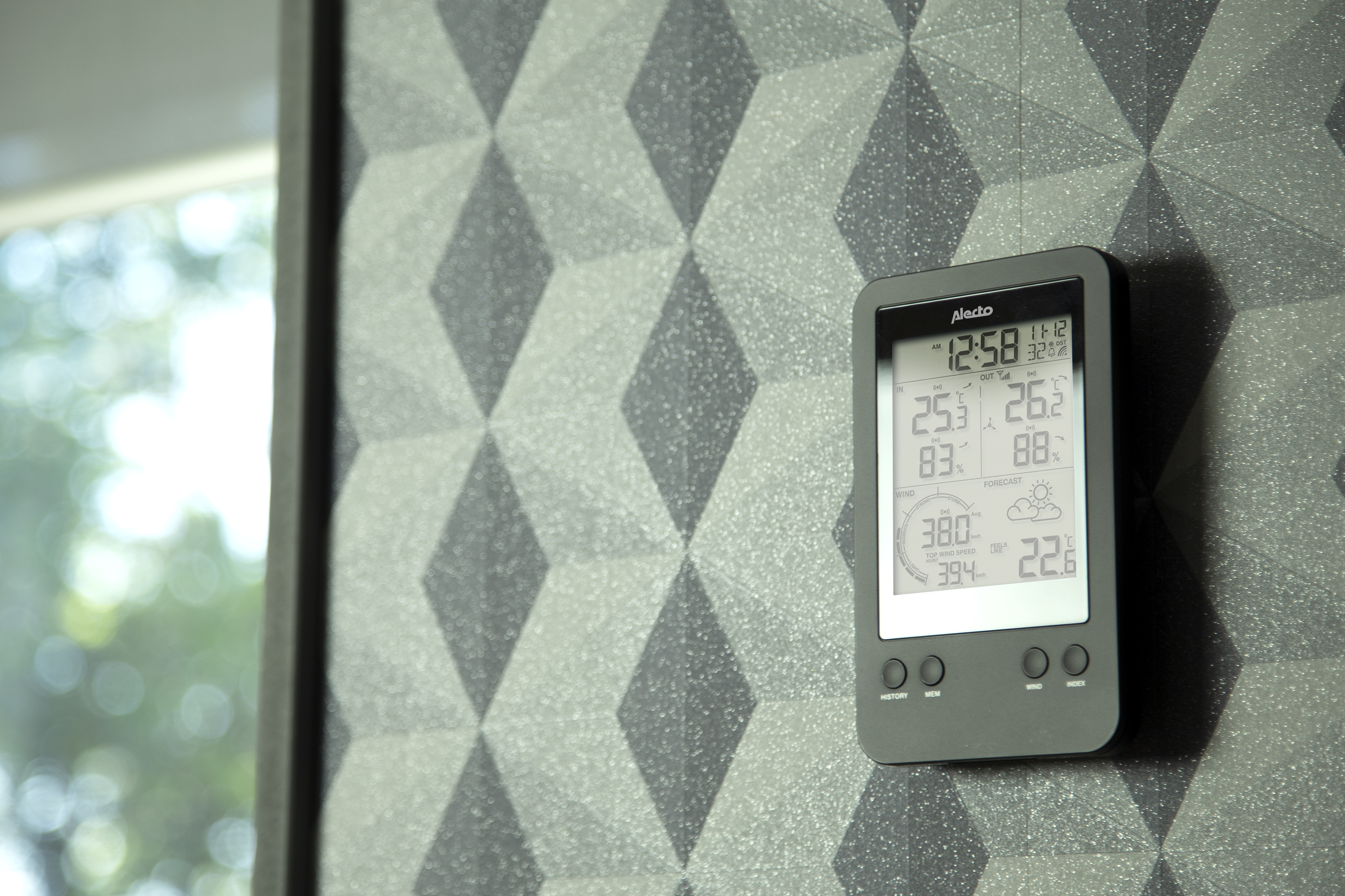 Cyfrowa stacja pogodowa na geometrycznej ścianie. Pokazuje czas, temp. wewnątrz/na zewnątrz, wilgotność i prognozę.