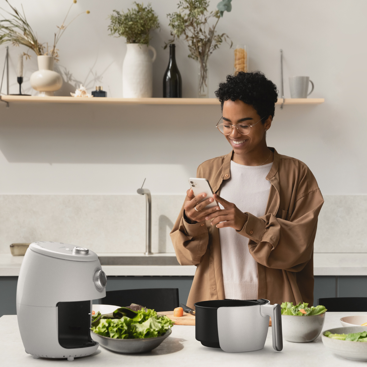 Mujer en la cocina usando un teléfono inteligente. Freidora de aire y ensalada en la encimera. Fondo blanco.