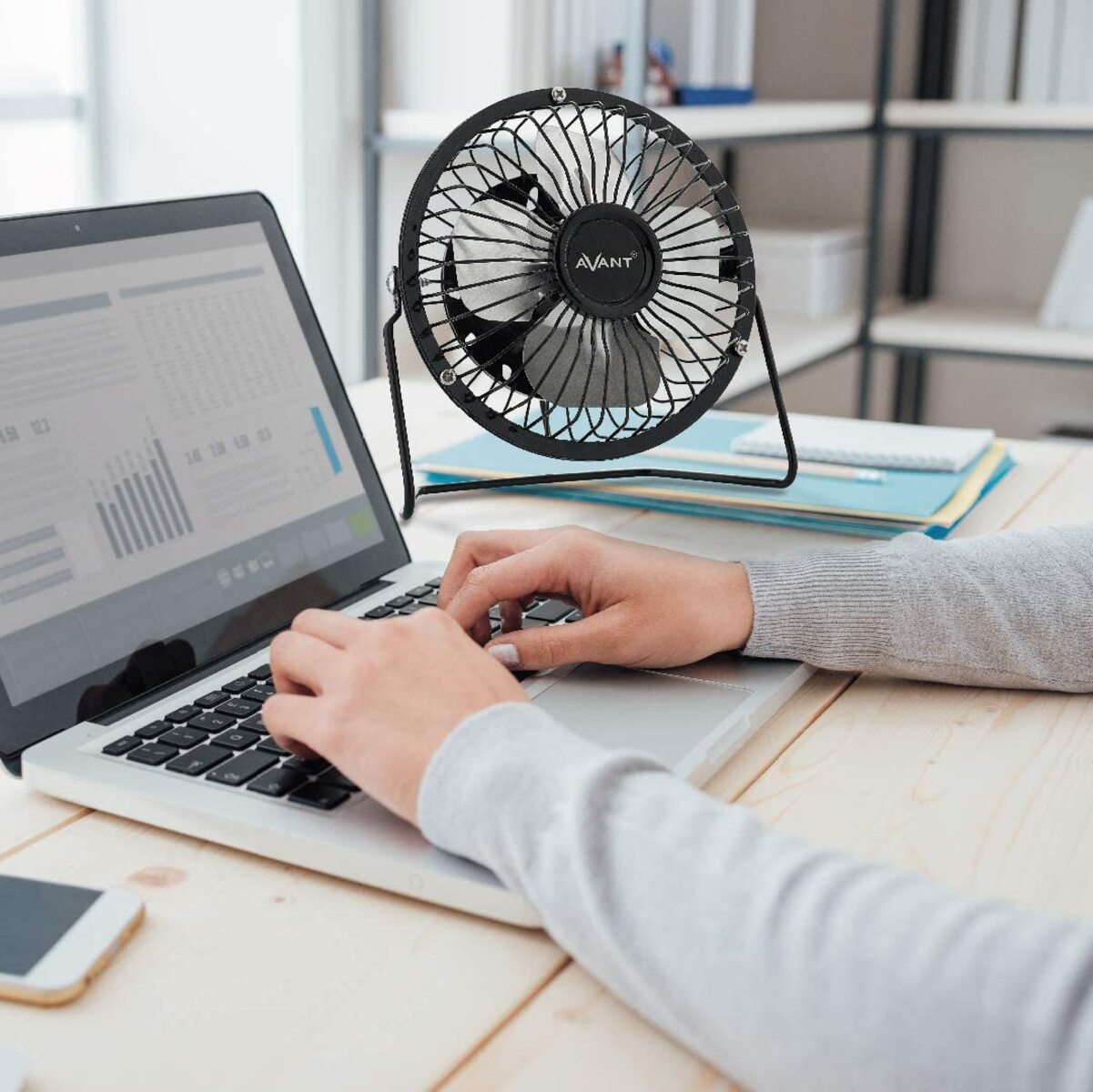 Persona escribiendo en un portátil con un ventilador de escritorio negro en el fondo. El ventilador tiene un marco negro.