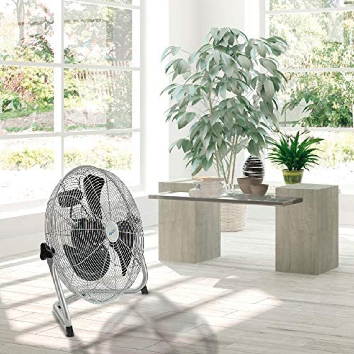 Un ventilatore d'argento è in una stanza con piante e un tavolo. Le finestre sono sullo sfondo.