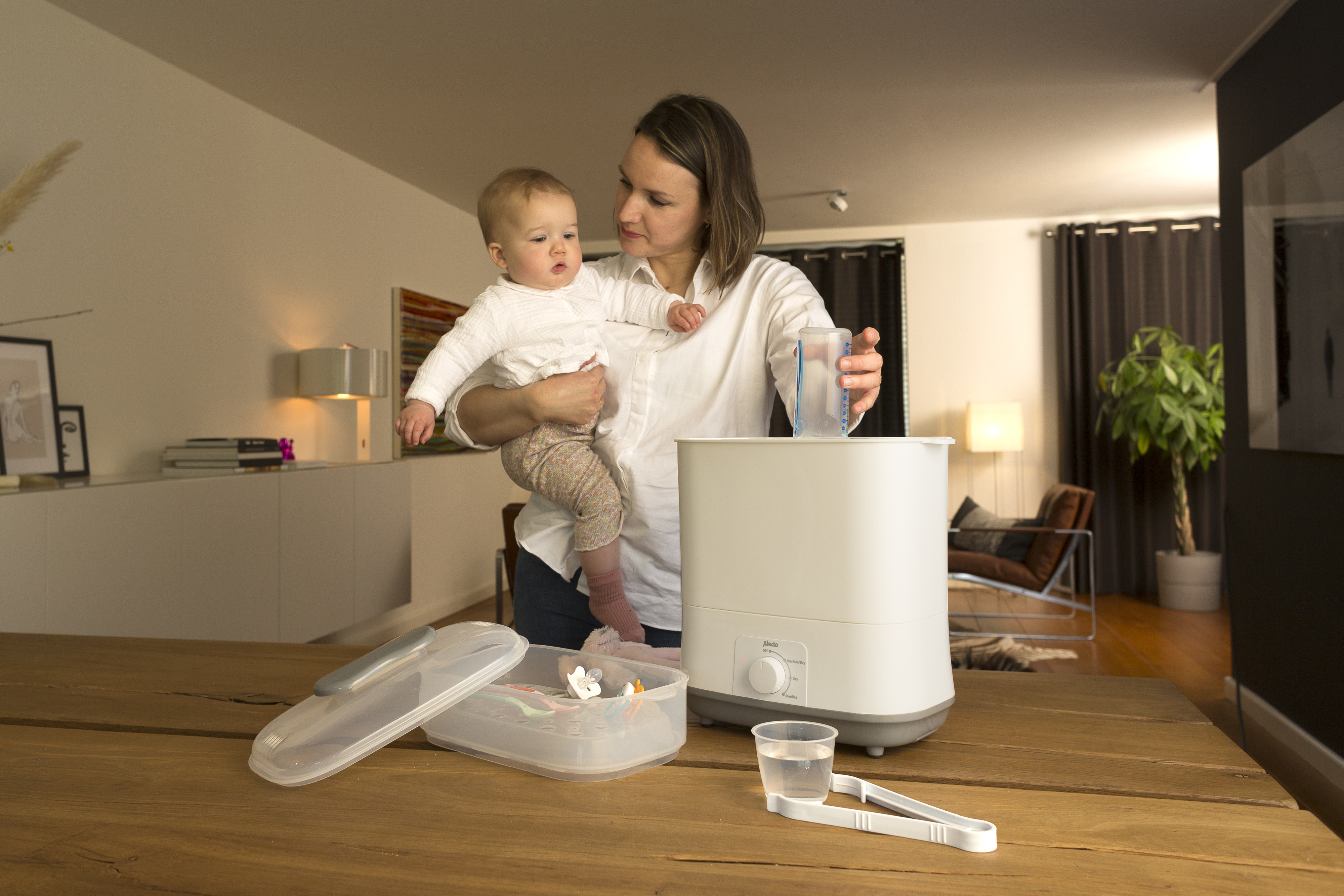 A woman holding a baby, preparing baby bottles. A sterilizer and other baby items are on the table.