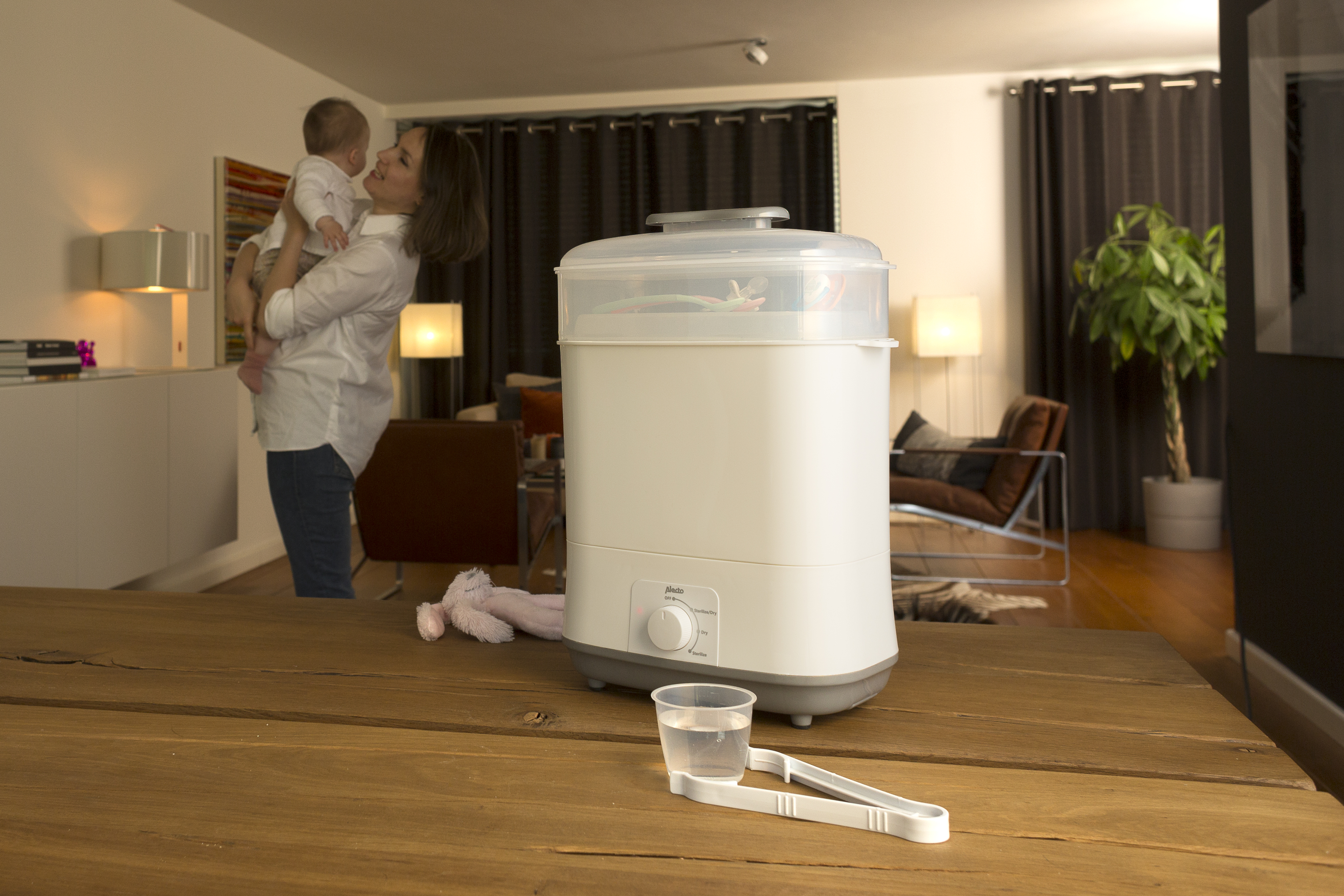 A woman holding a baby, with a baby bottle sterilizer and accessories on a table.