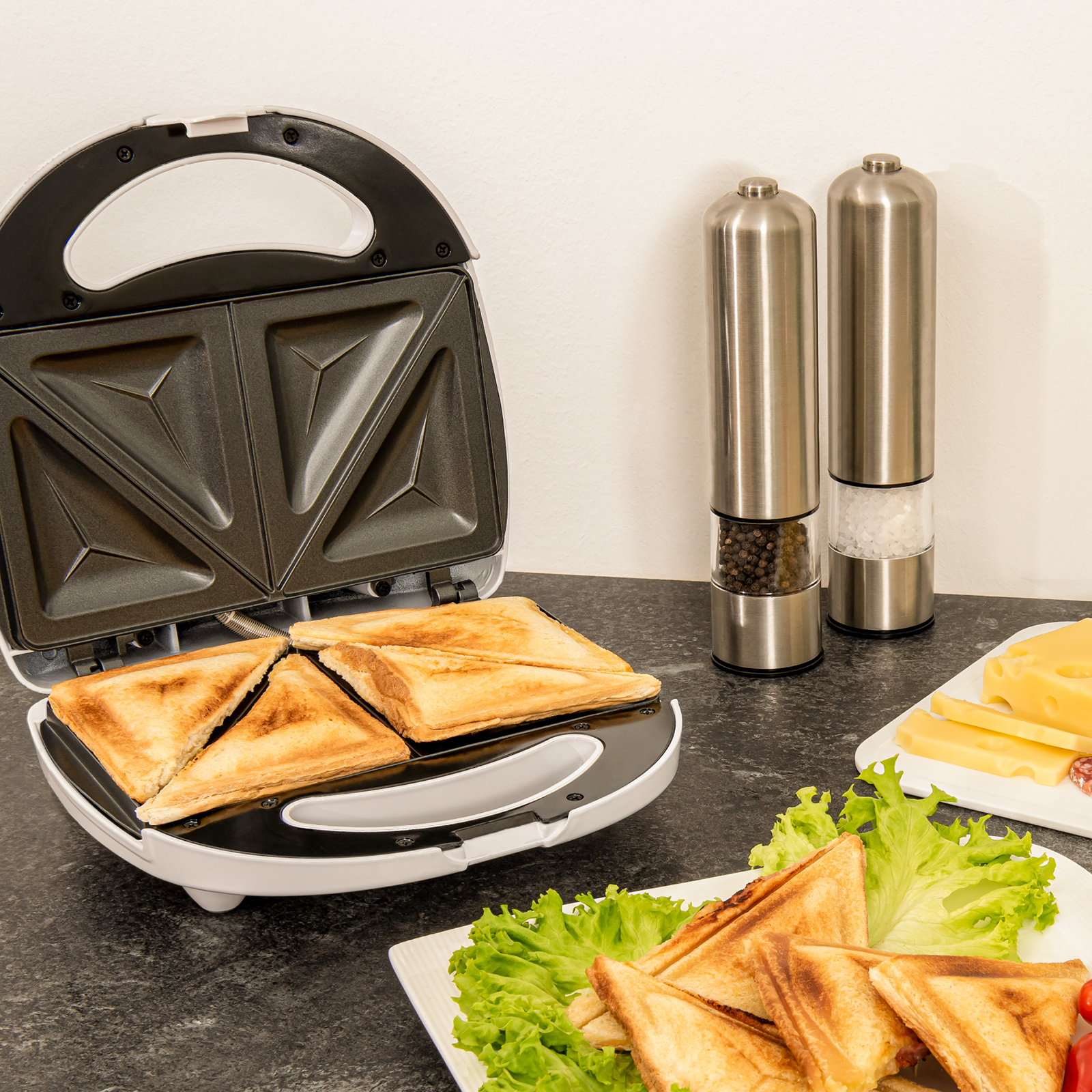 Ein Sandwichmaker mit Sandwiches. Pfeffer- und Salzstreuer stehen rechts. Käse und Salat sind in der Nähe.