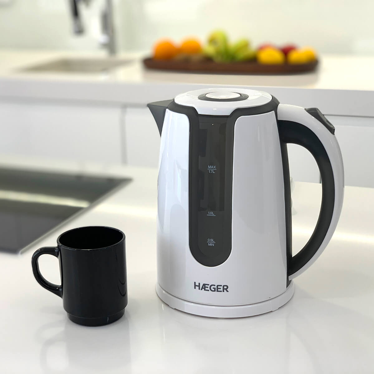 Hervidor eléctrico blanco y una taza negra sobre encimera blanca. Fondo borroso con comida visible.